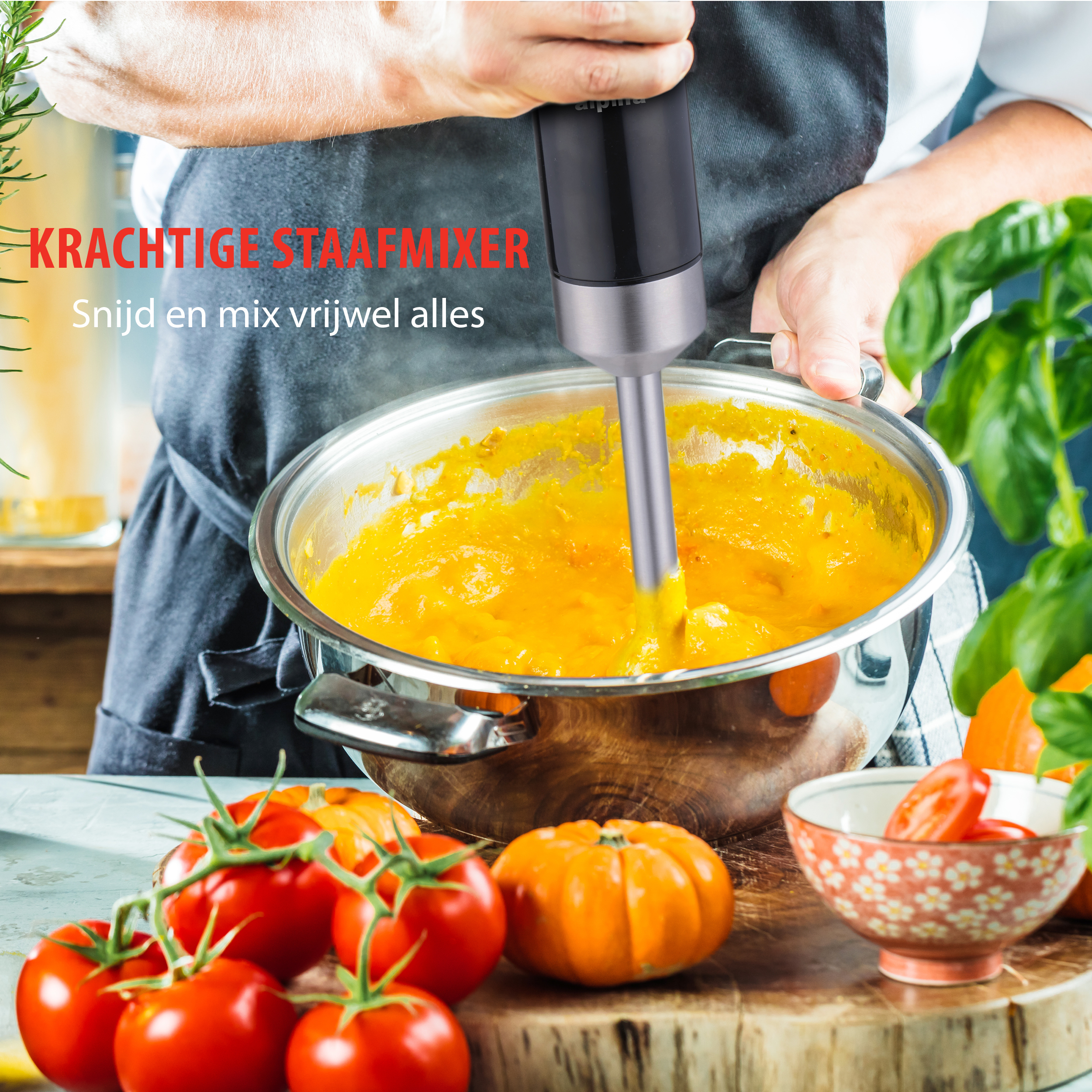 Persoon gebruikt een zwarte staafmixer in een pan met oranje soep. Tomaten en pompoen op een plank.
