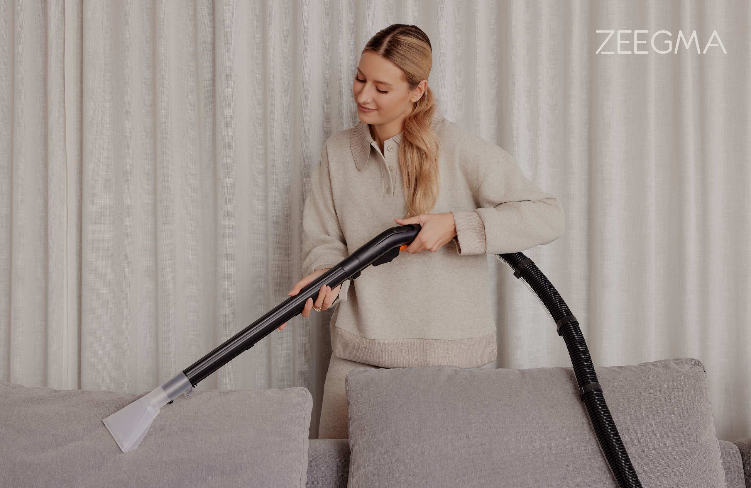 Kobieta odkurza szarą sofę odkurzaczem ręcznym. Beżowy strój.