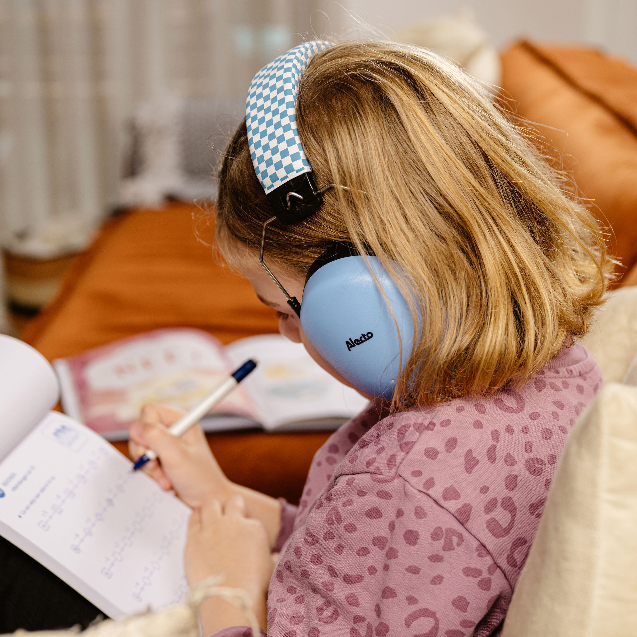 Dziewczyna w słuchawkach pisząca w notatniku. Pomarańczowa kanapa i książki w tle.