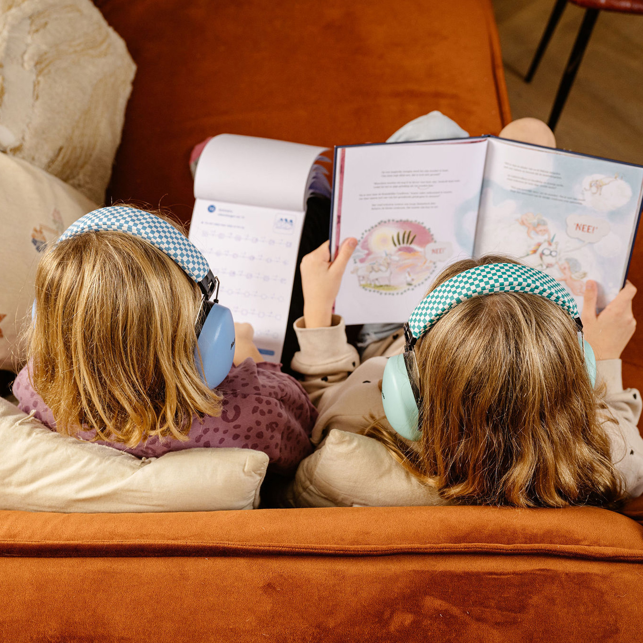 Dwoje dzieci czyta razem książkę, mając na sobie słuchawki z redukcją szumów. Siedzą na pomarańczowej kanapie.