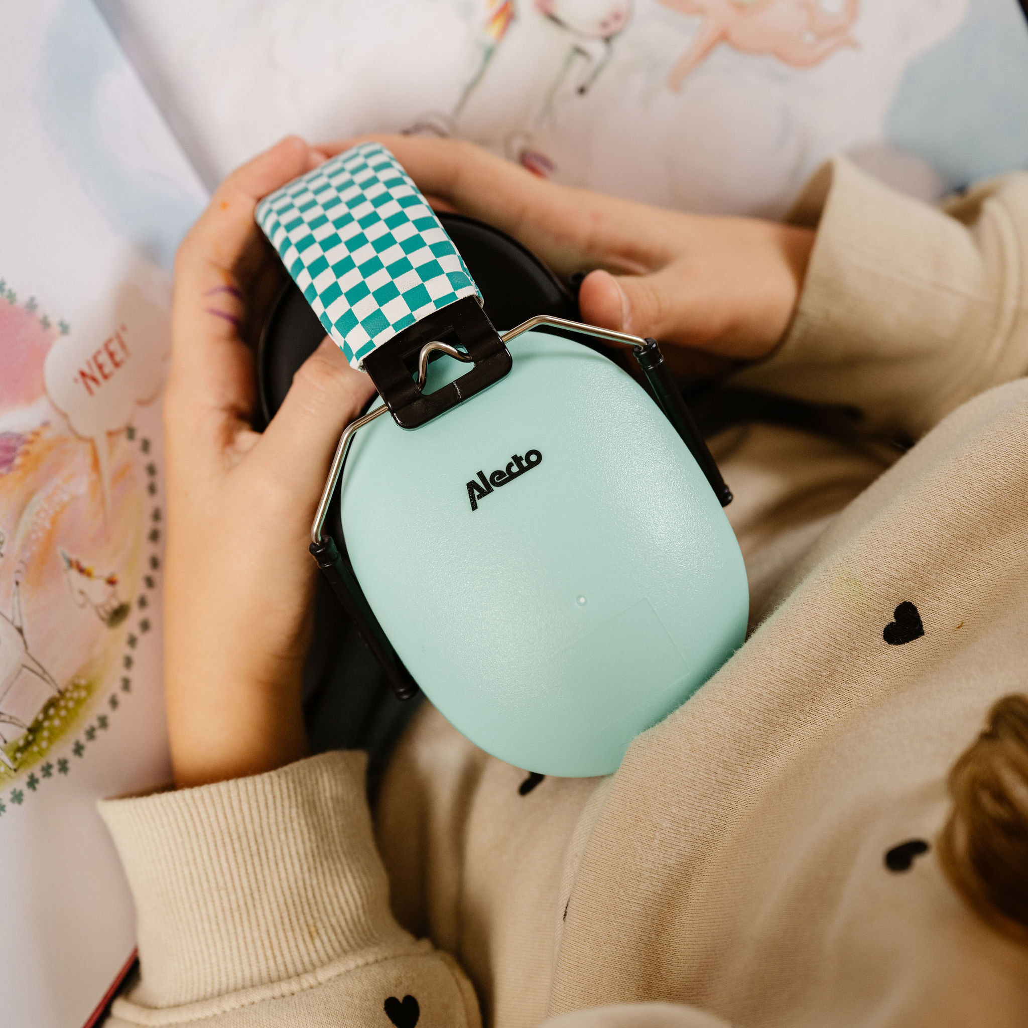 Ręce dziecka trzymają miętowo-zielone nauszniki z szachownicą. Na nausznikach napis "Alecto".