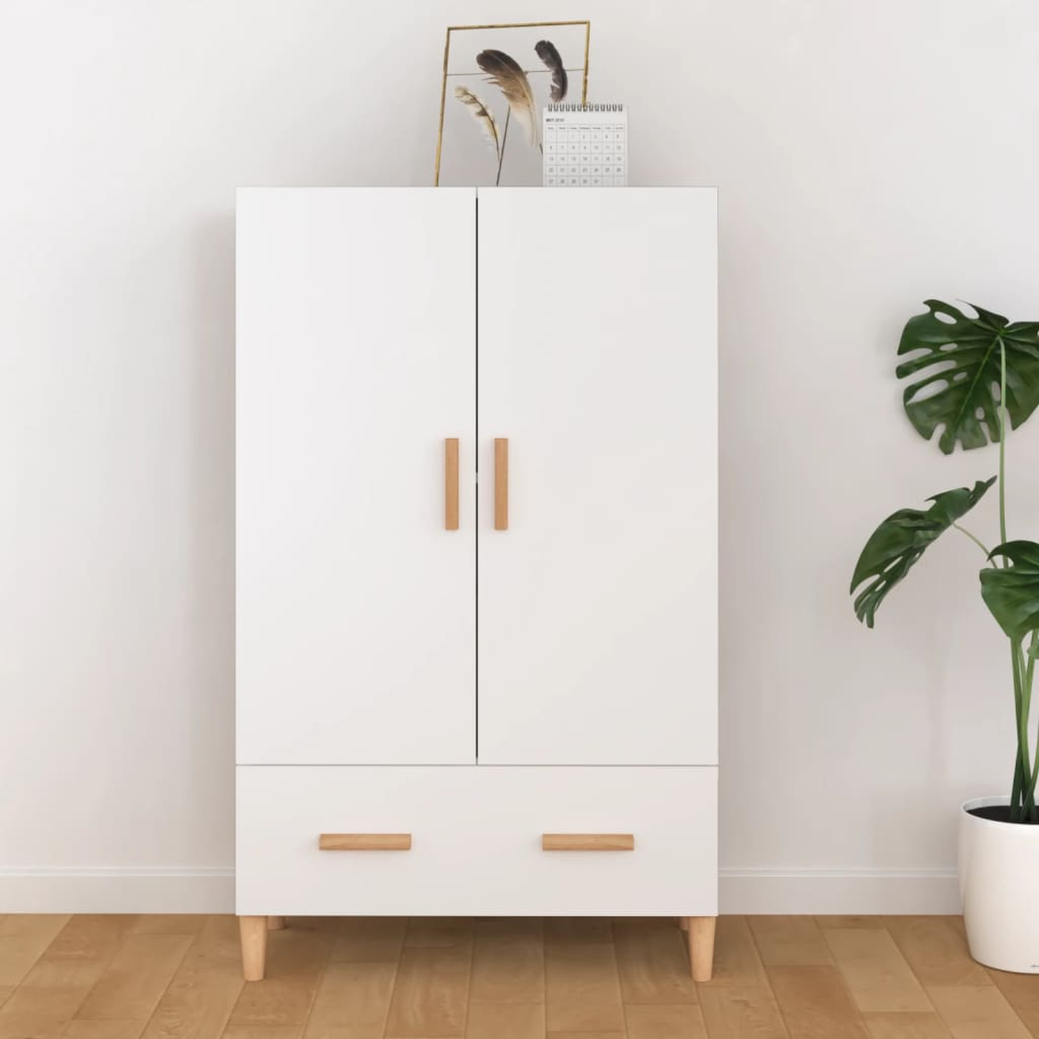 Weißer Kleiderschrank mit zwei Türen und einer Schublade. Holzgriffe und -beine. Pflanze und Kunstwerk daneben.