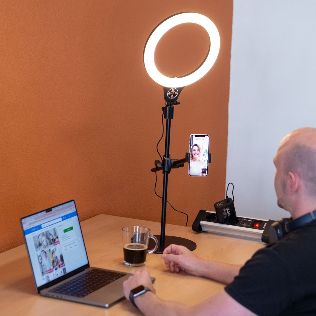 Een man die een ringlicht en een laptop gebruikt. Telefoon op standaard, koffiekopje op tafel.