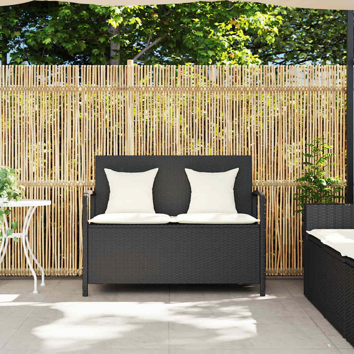 Divano nero in vimini con due cuscini bianchi, davanti a una recinzione di bambù. Un tavolino a sinistra.