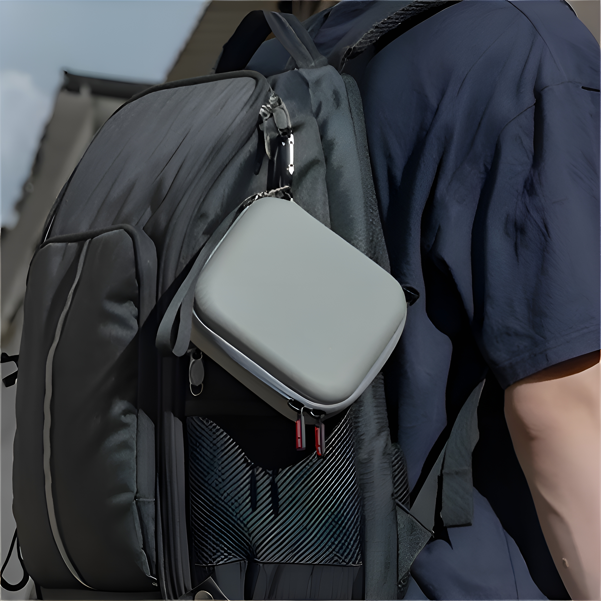 Eine Person mit Rucksack hält einen grauen quadratischen Koffer mit Reißverschluss und kleinem Riemen.