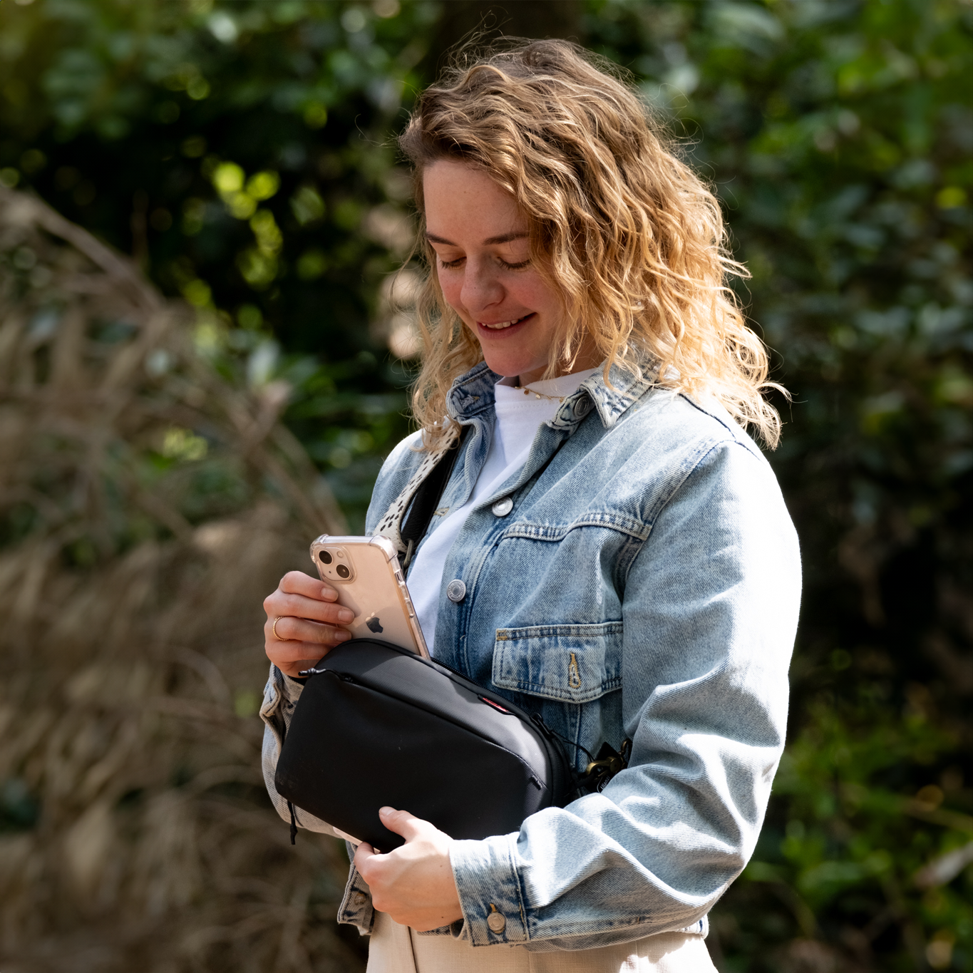 Kobieta trzymająca telefon w czarnej torbie, uśmiechnięta w naturalnym otoczeniu.