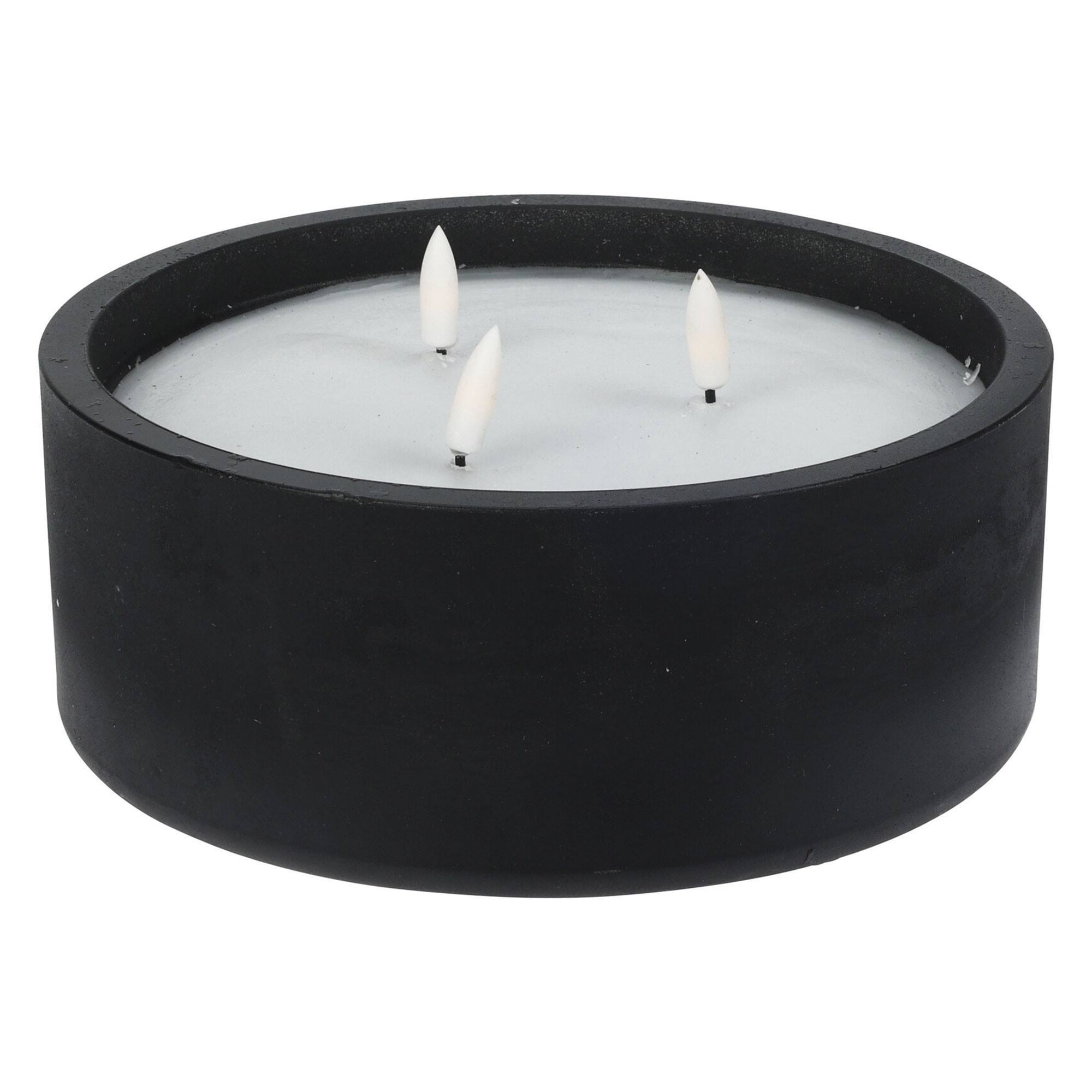 Vela negra con tres llamas blancas. La vela está en un recipiente negro y redondo sobre una superficie blanca.