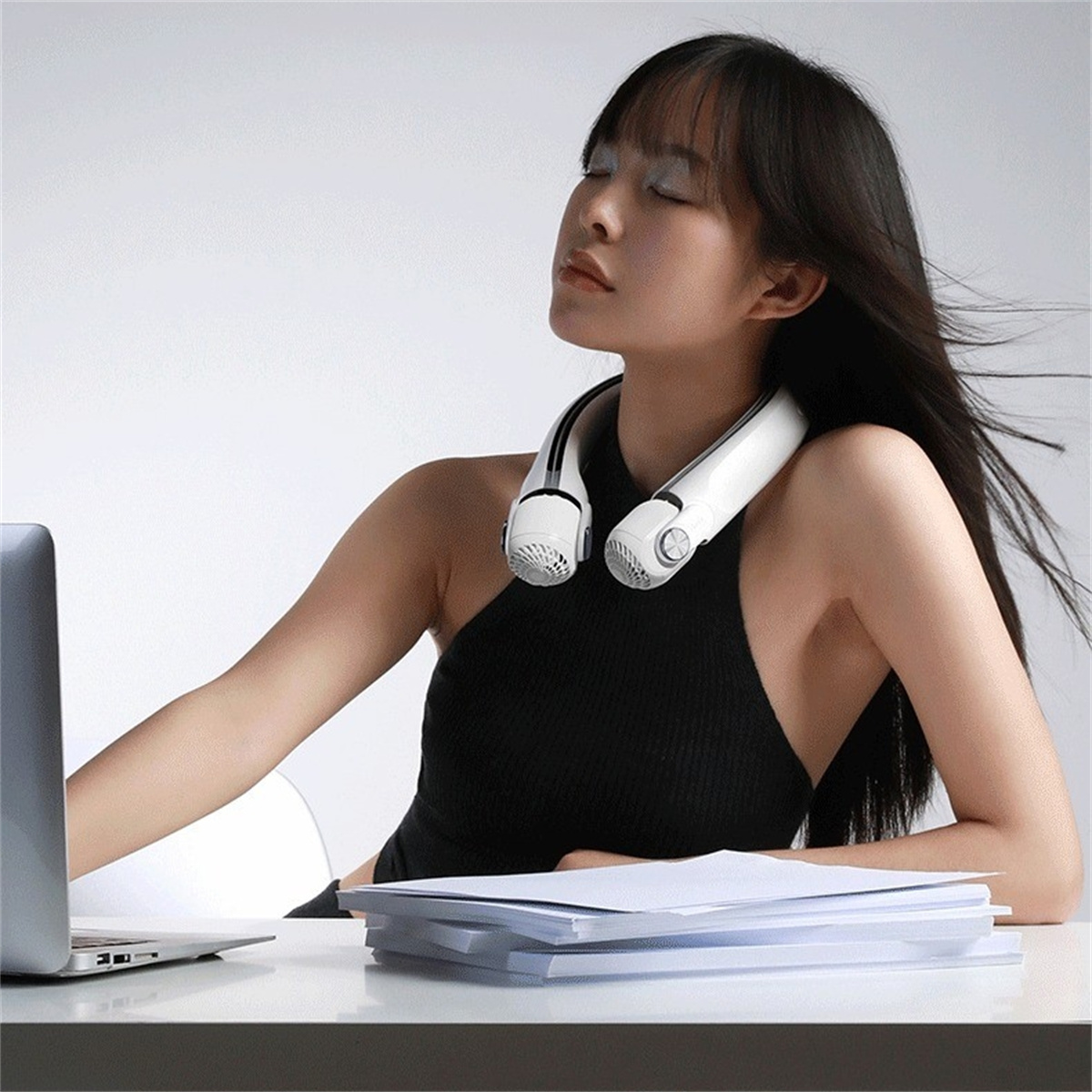Eine Frau, die einen weißen Nackenfächer trägt, arbeitet an einem Schreibtisch mit Laptop und Papieren, Augen geschlossen.