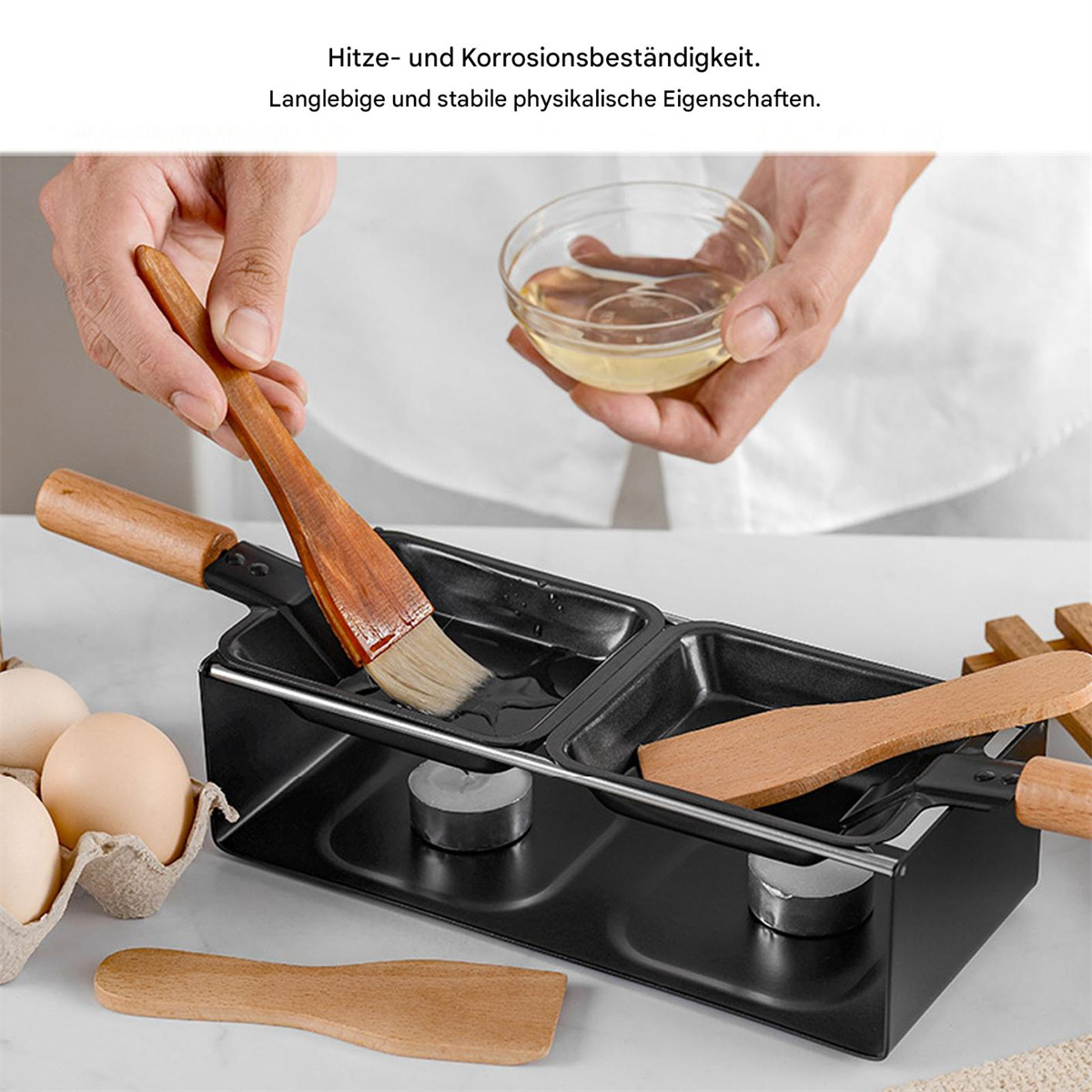 Ein schwarzer Grill mit Holzgriffen und eine Schüssel mit Öl, gehalten von Händen, und Eier im Hintergrund.