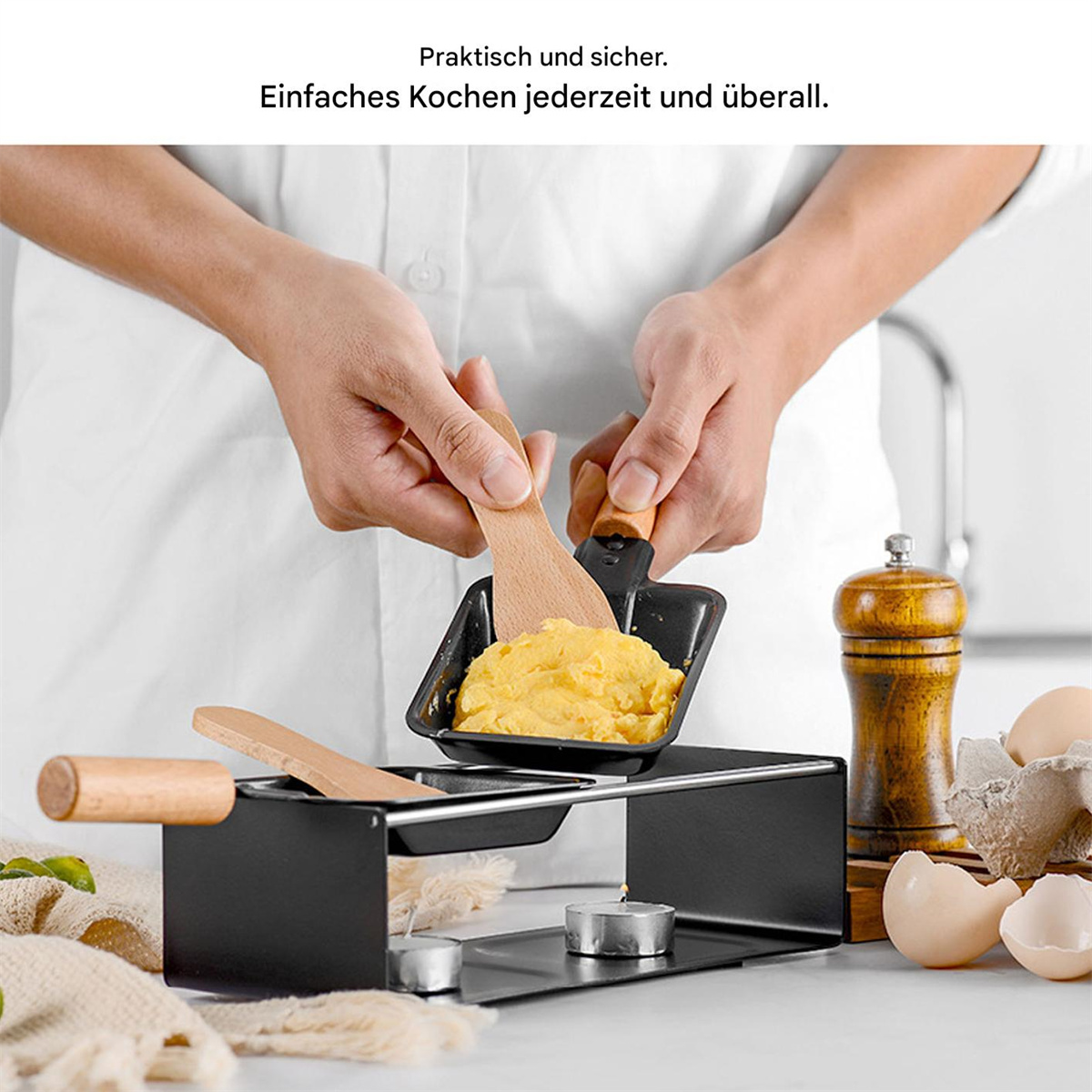 Hände halten Raclette-Pfanne mit geschmolzenem Käse, Holzspateln und Eiern.