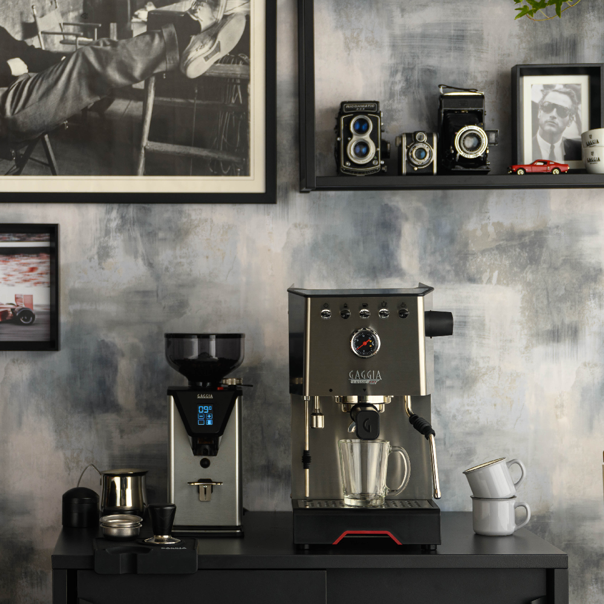 Eine Kaffeemaschine und eine Mühle auf einem schwarzen Regal. Andere Gegenstände sind vorhanden, wie Kaffeetassen und Fotos.
