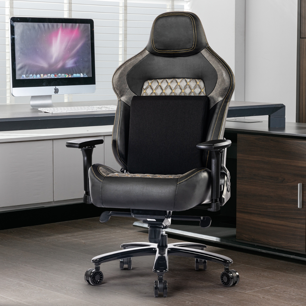 Schwarzer und grauer Gaming-Stuhl mit Lendenwirbelstütze vor einem Schreibtisch mit Computer und Fenster mit Jalousien.