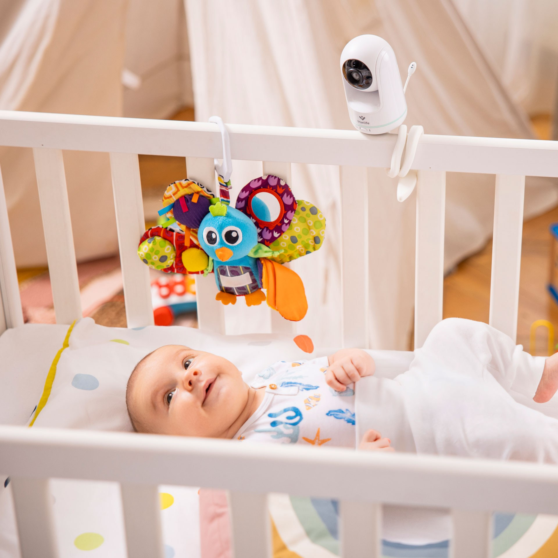 Baby im Kinderbett mit Mobile und Kamera, weißes Bett und Hintergrund.