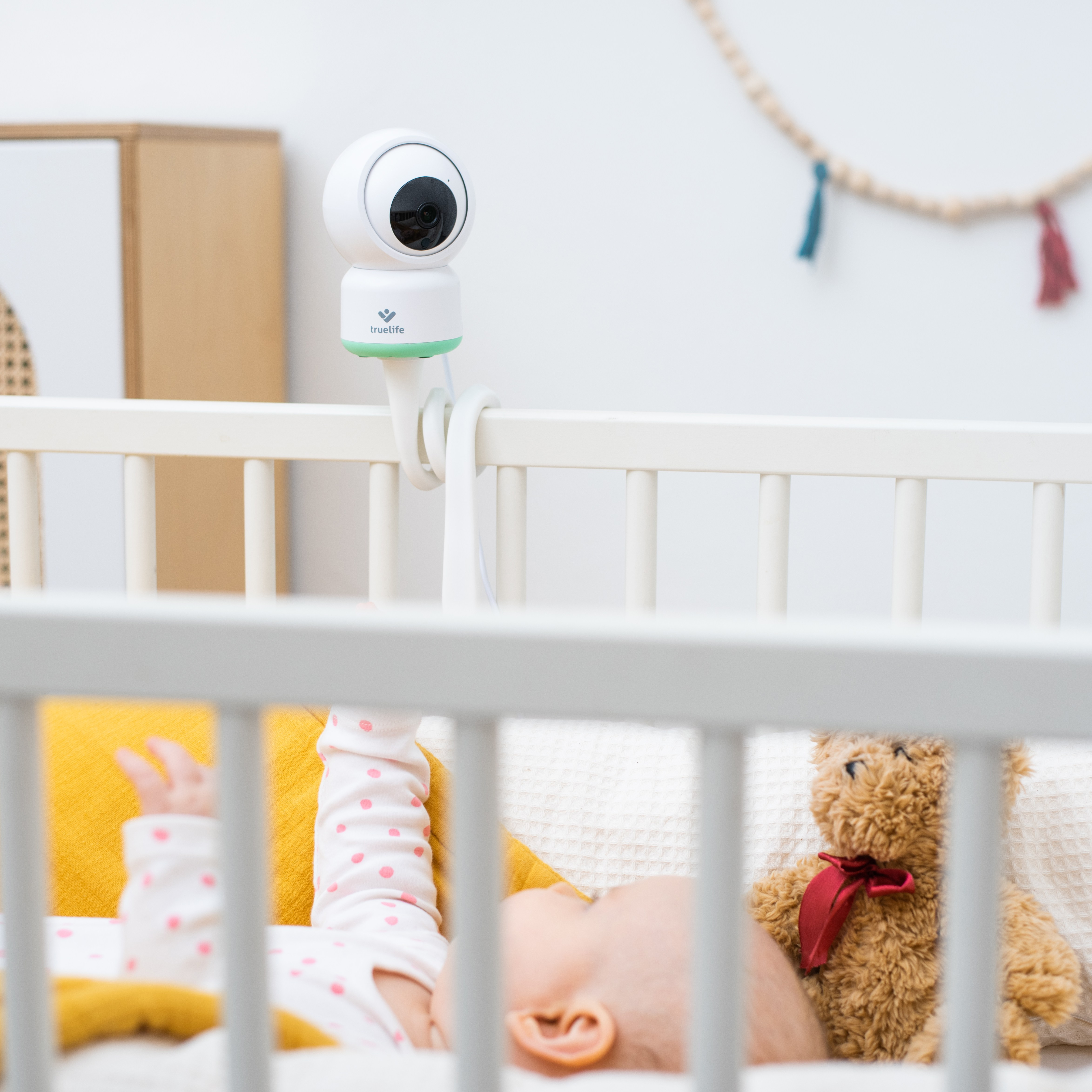 Ein Babyphone mit einem Baby in einem Kinderbett. Ein Teddybär ist in der Nähe des Babys, in einem gut beleuchteten Raum.