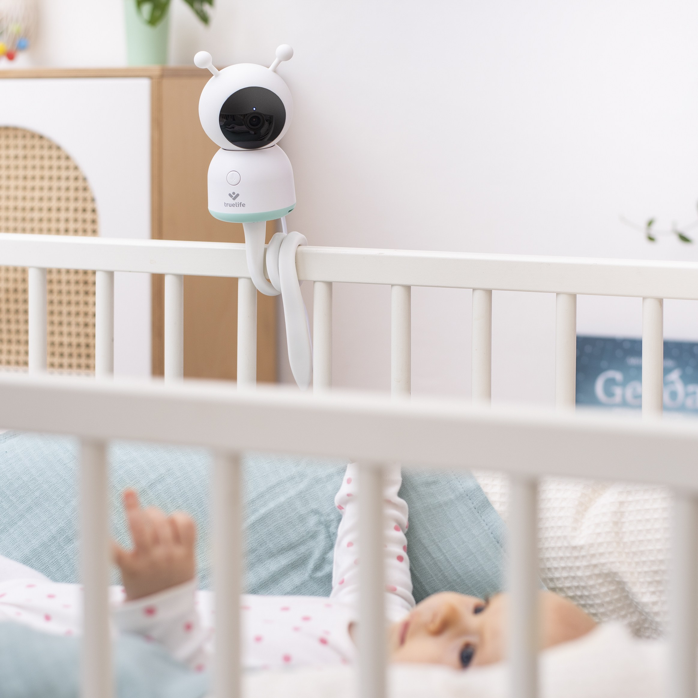 Babyphone an einem Gitterbett befestigt. Ein Baby liegt im Bett. Der Monitor ist weiß und blau.