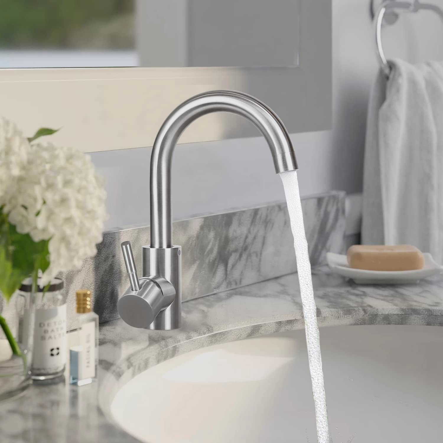 Ein verchromter Wasserhahn mit Wasser, das in ein weißes Waschbecken fließt. Seife, Blumen und Flaschen sind sichtbar.