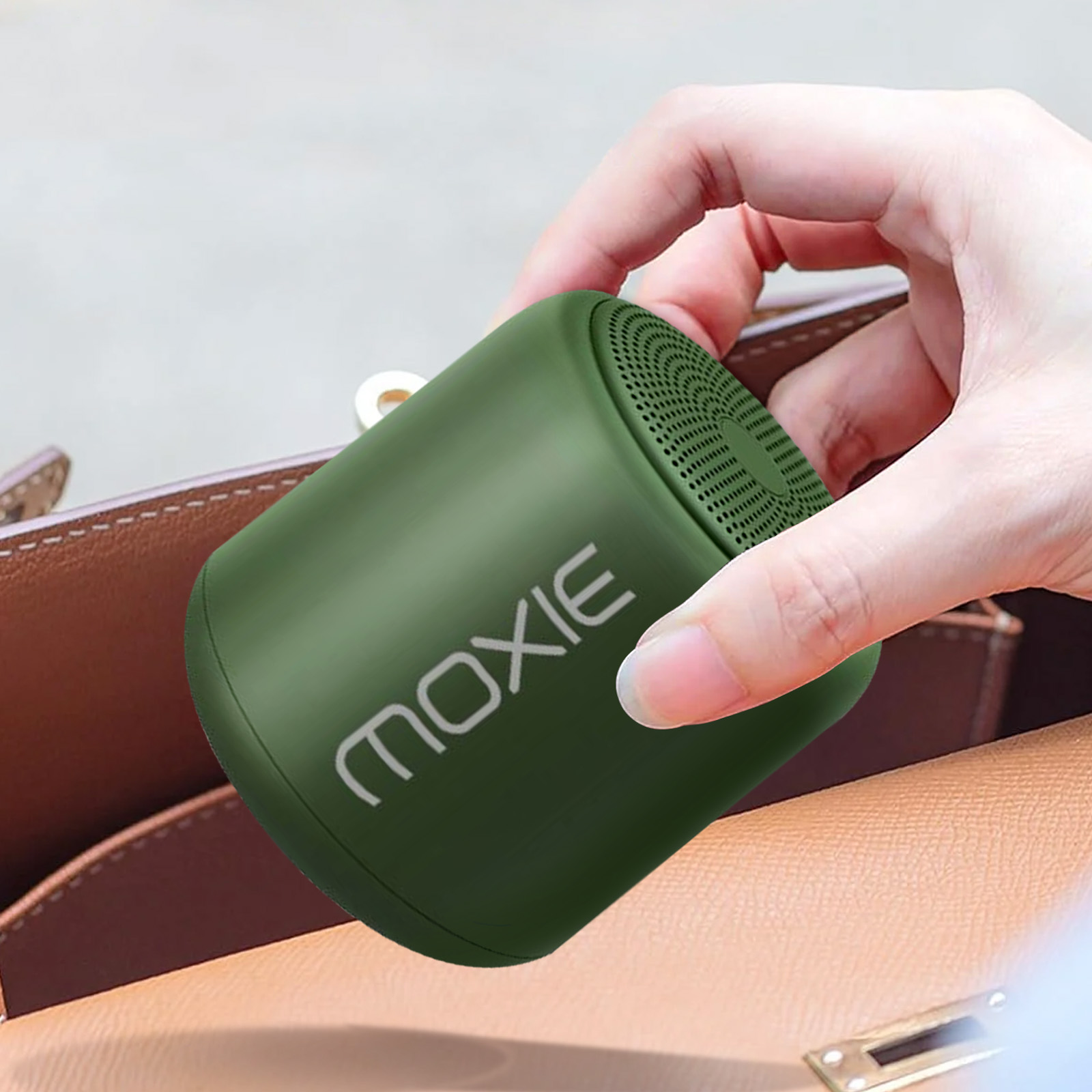 Eine Hand hält einen grünen Lautsprecher aus einer braunen Handtasche. Der Lautsprecher hat das Wort MOXIE.