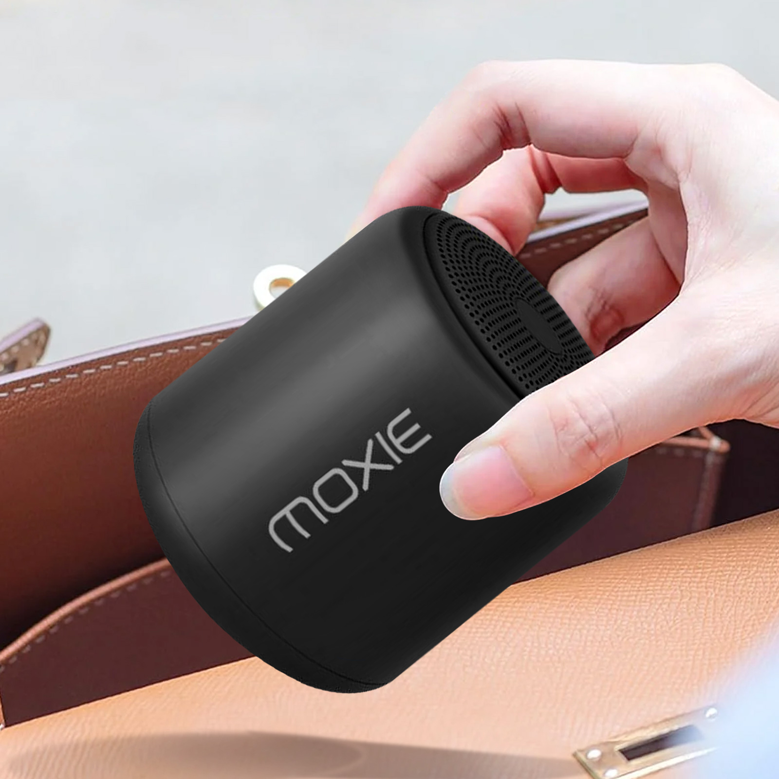 Un petit haut-parleur noir étiqueté 'moxie' est tenu dans une main, à côté d'un sac à main marron.