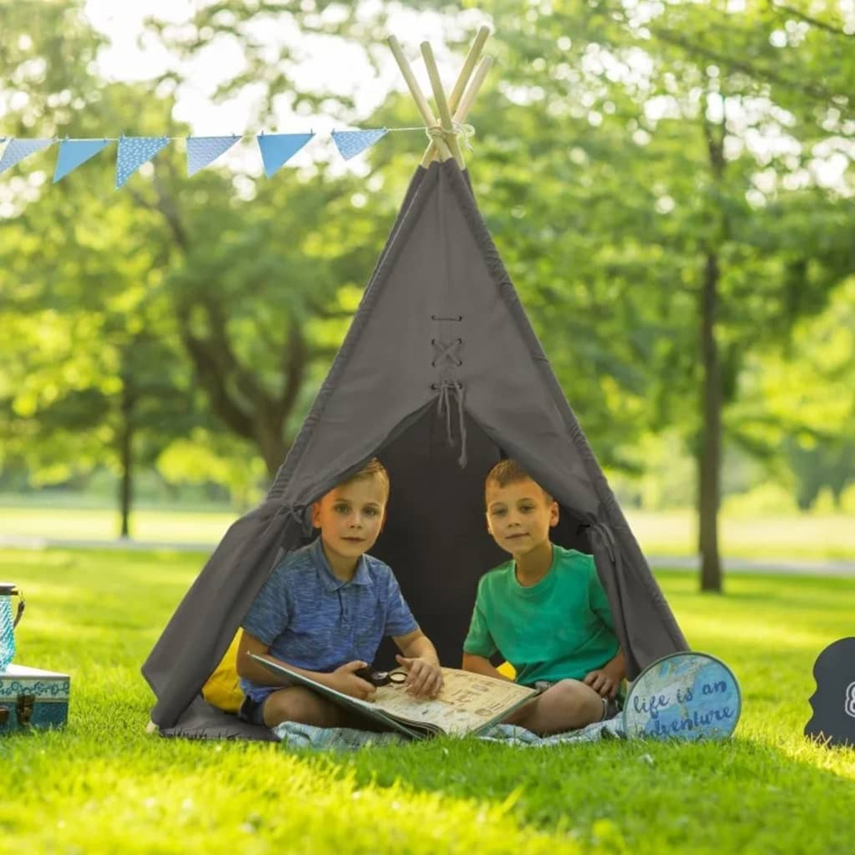 Zwei Jungen sitzen in einem grauen Tipi-Zelt in einem Park. Sie schauen sich ein Buch an.