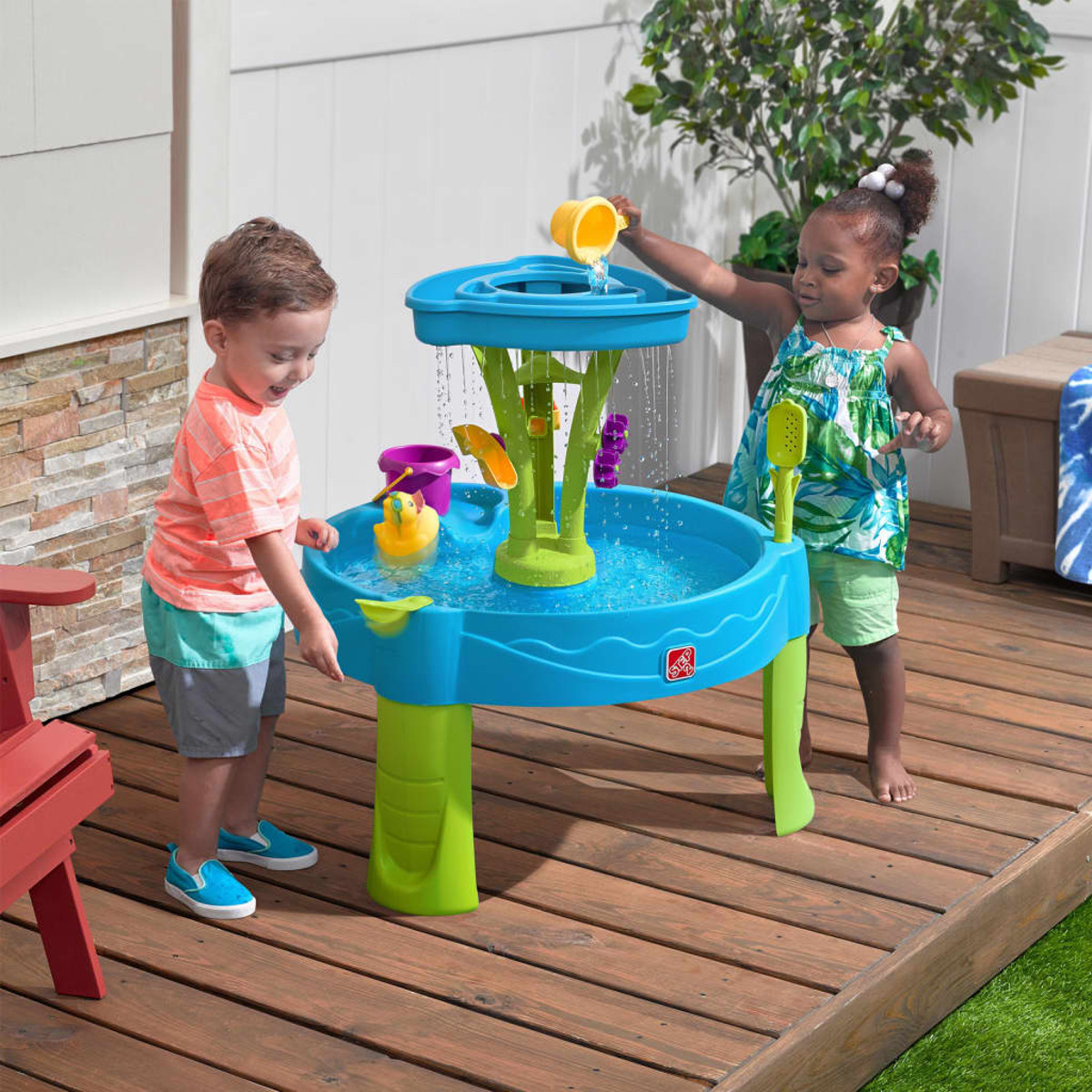 Zwei Kinder spielen mit einem Wassertisch. Es gibt einen blau-grünen Tisch mit Wasser und Spielzeug.