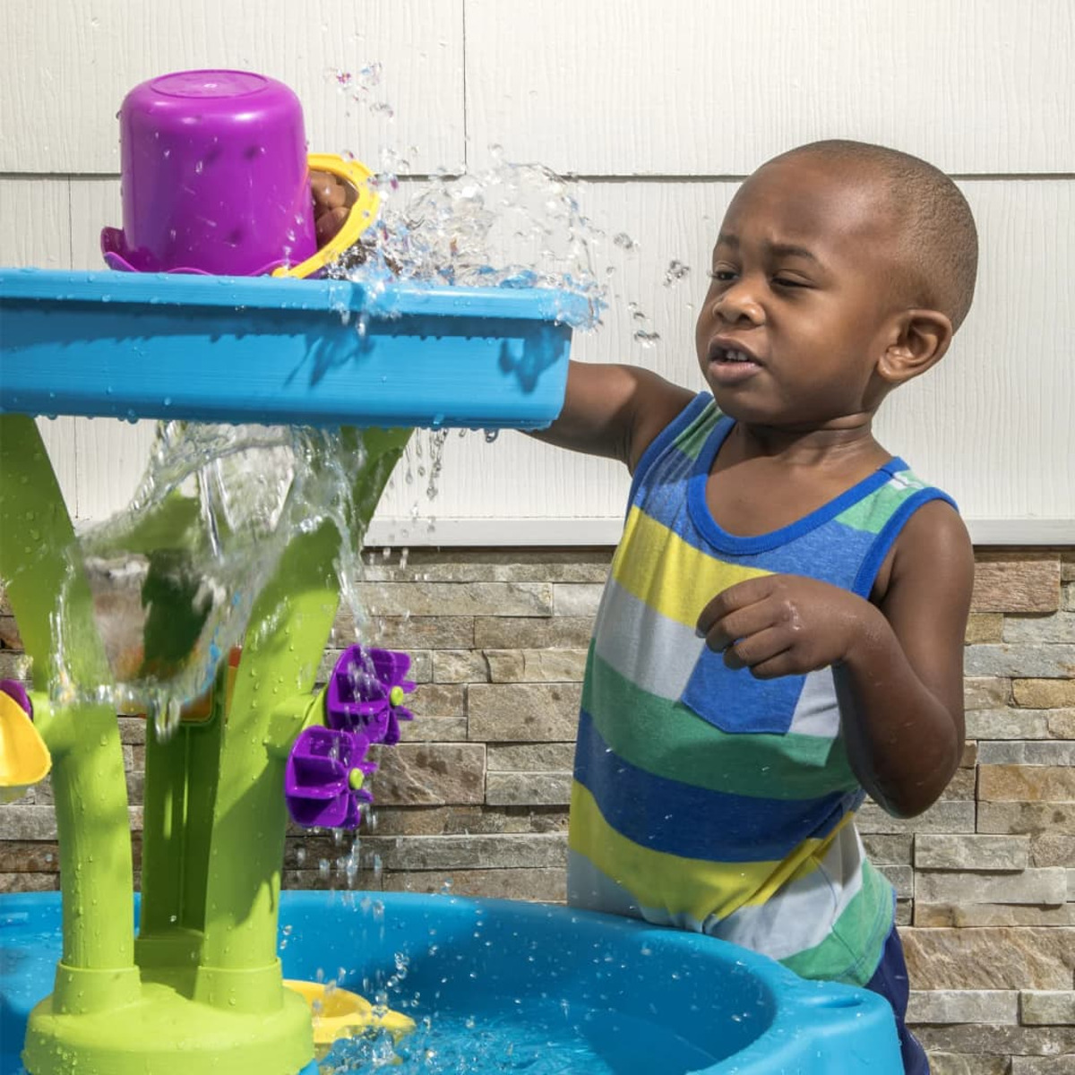 Ein Kleinkind spielt mit einem Wassertisch. Wasser spritzt. Er trägt ein gestreiftes Hemd.