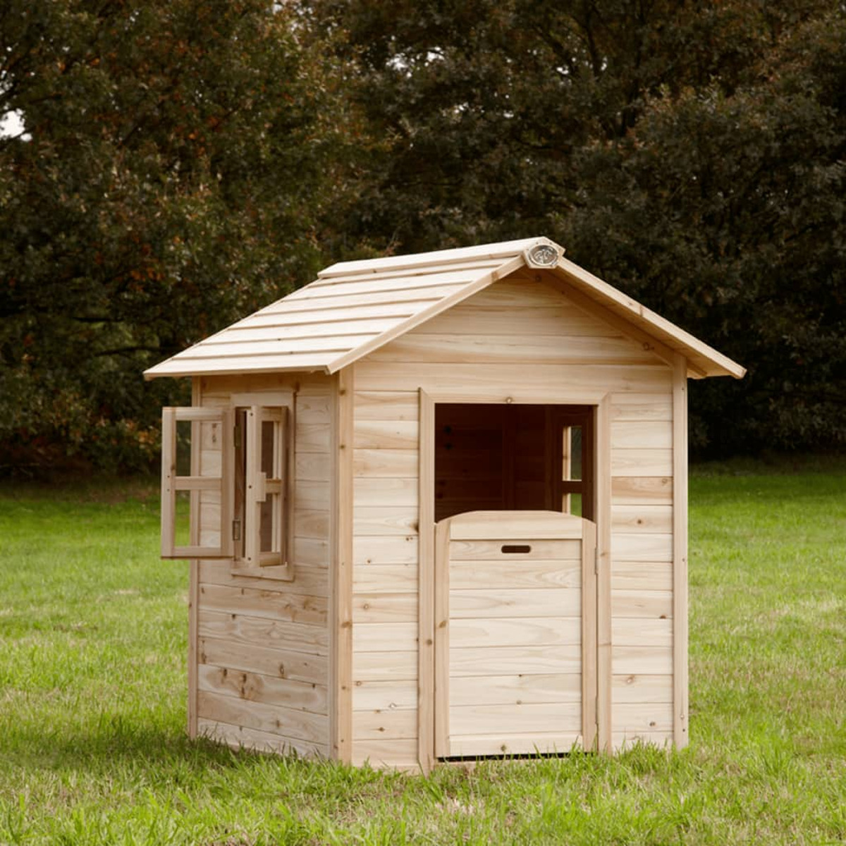Holzspielhaus mit offener Tür und Fenster auf grünem Gras, Bäume im Hintergrund.