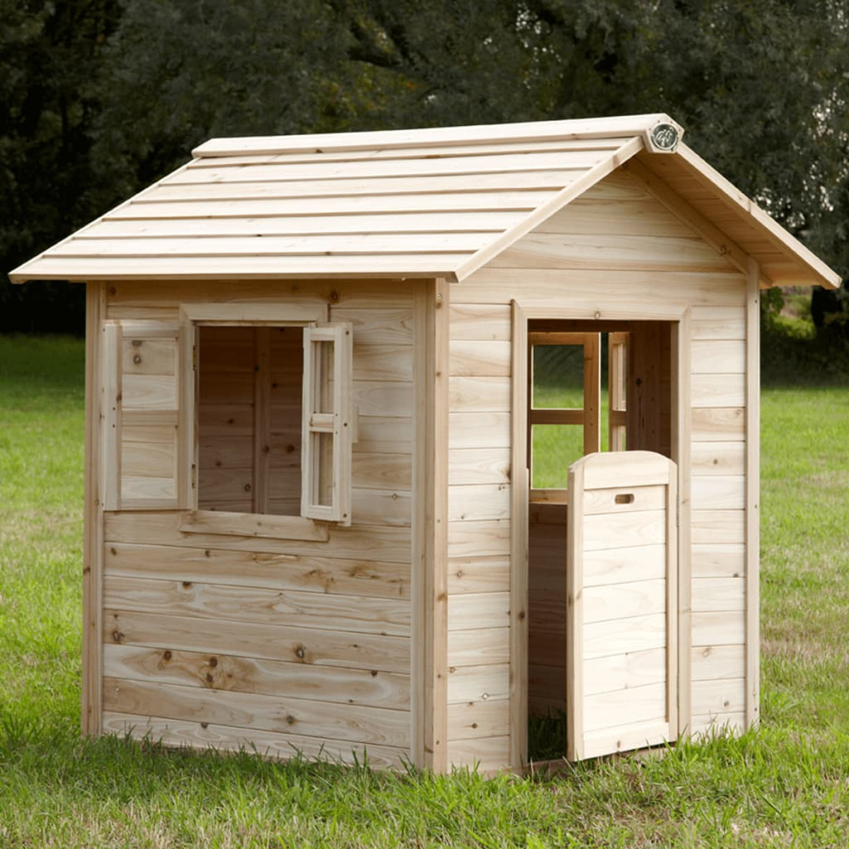 Ein Holzspielhaus auf grünem Gras. Es hat ein offenes Fenster und eine Tür. Im Hintergrund sind Bäume.