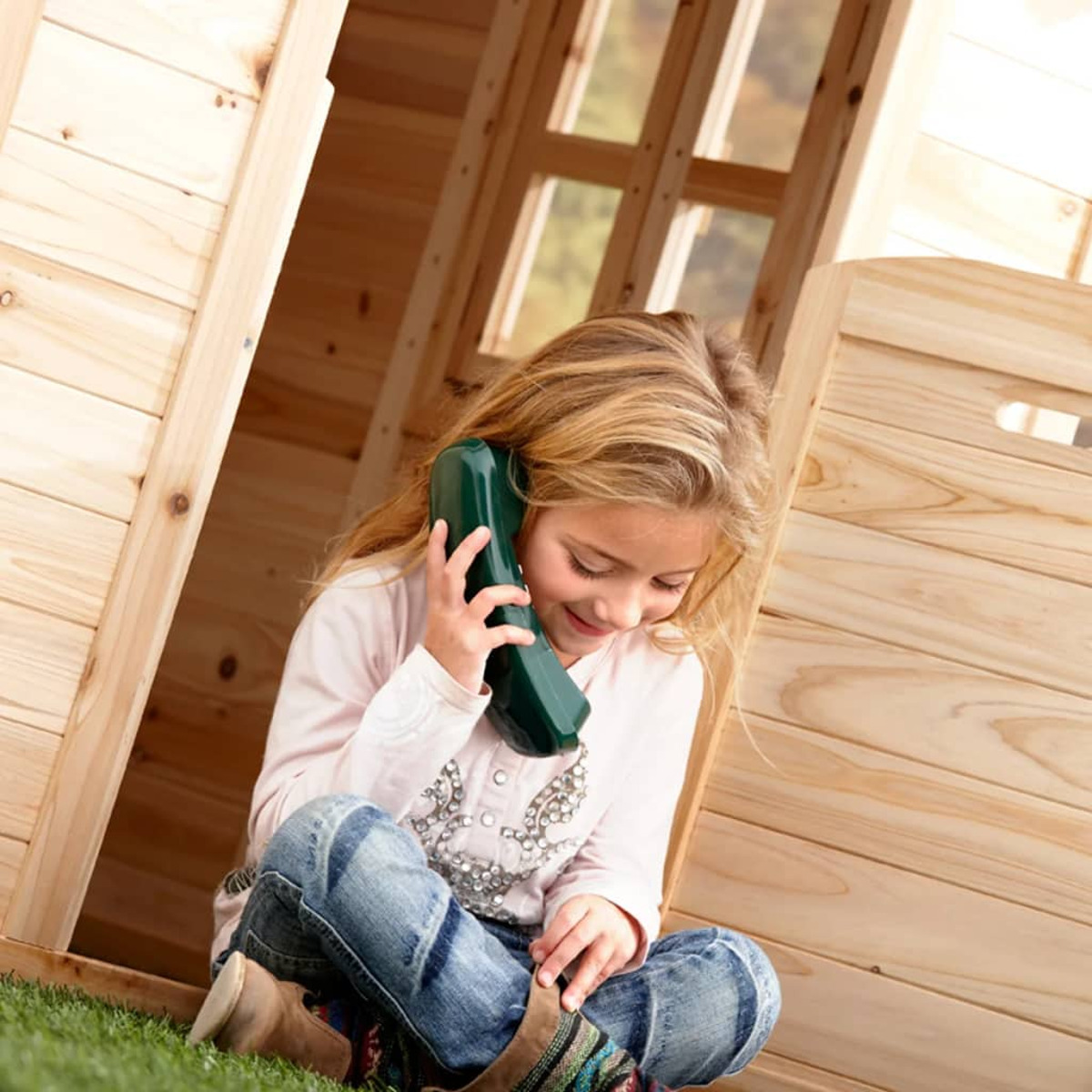 Ein Mädchen sitzt in einem Holzspielhaus und telefoniert mit einem grünen Spielzeugtelefon.