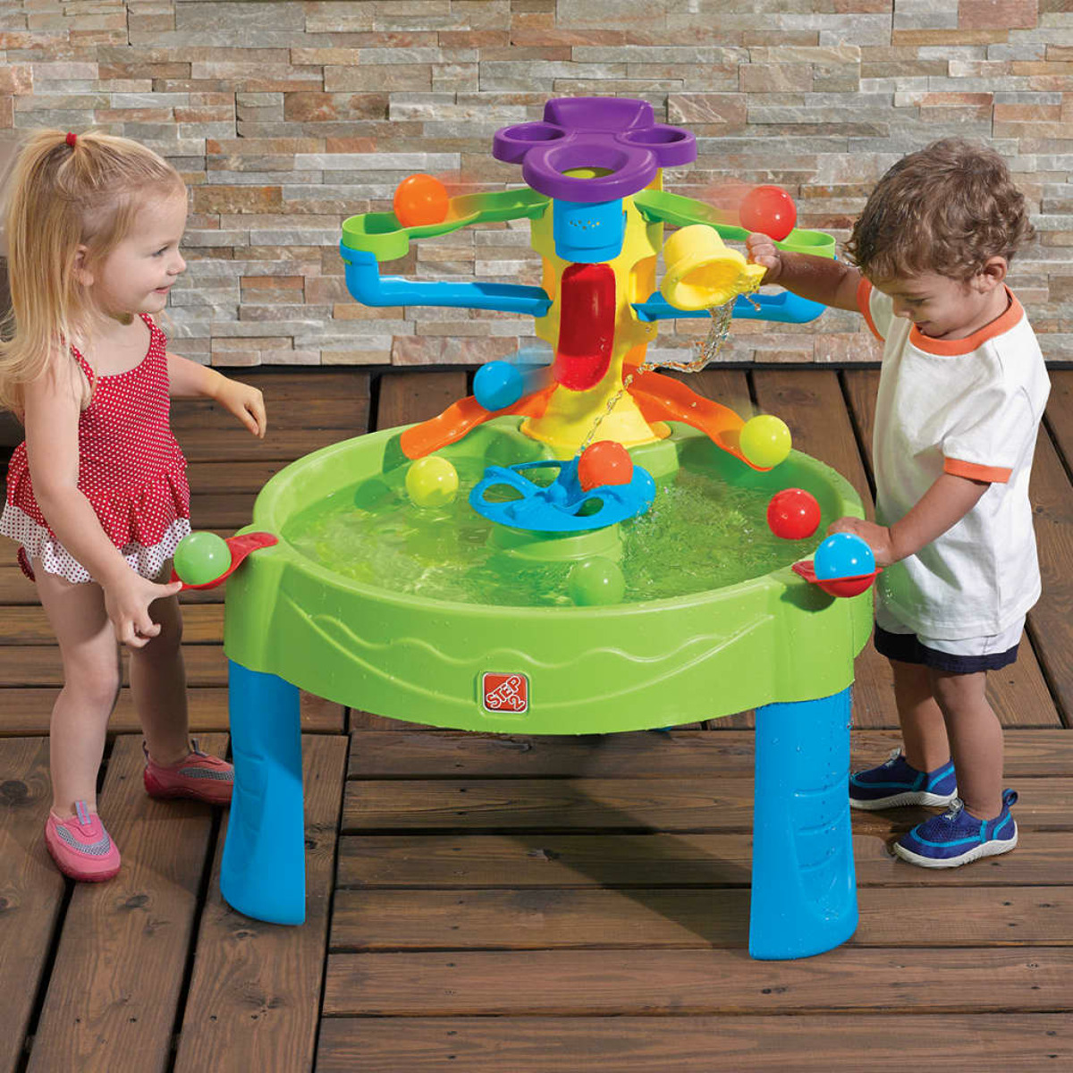 Zwei Kinder spielen mit einem Wassertisch. Grüner, blauer und lila Tisch. Hintergrund ist eine Ziegelmauer.