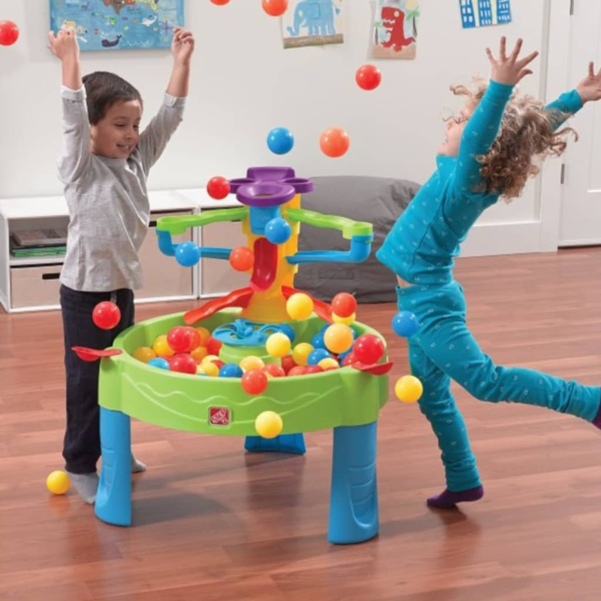 Zwei Kinder spielen mit einem Balltisch, umgeben von bunten Bällen, auf einem Holzboden.