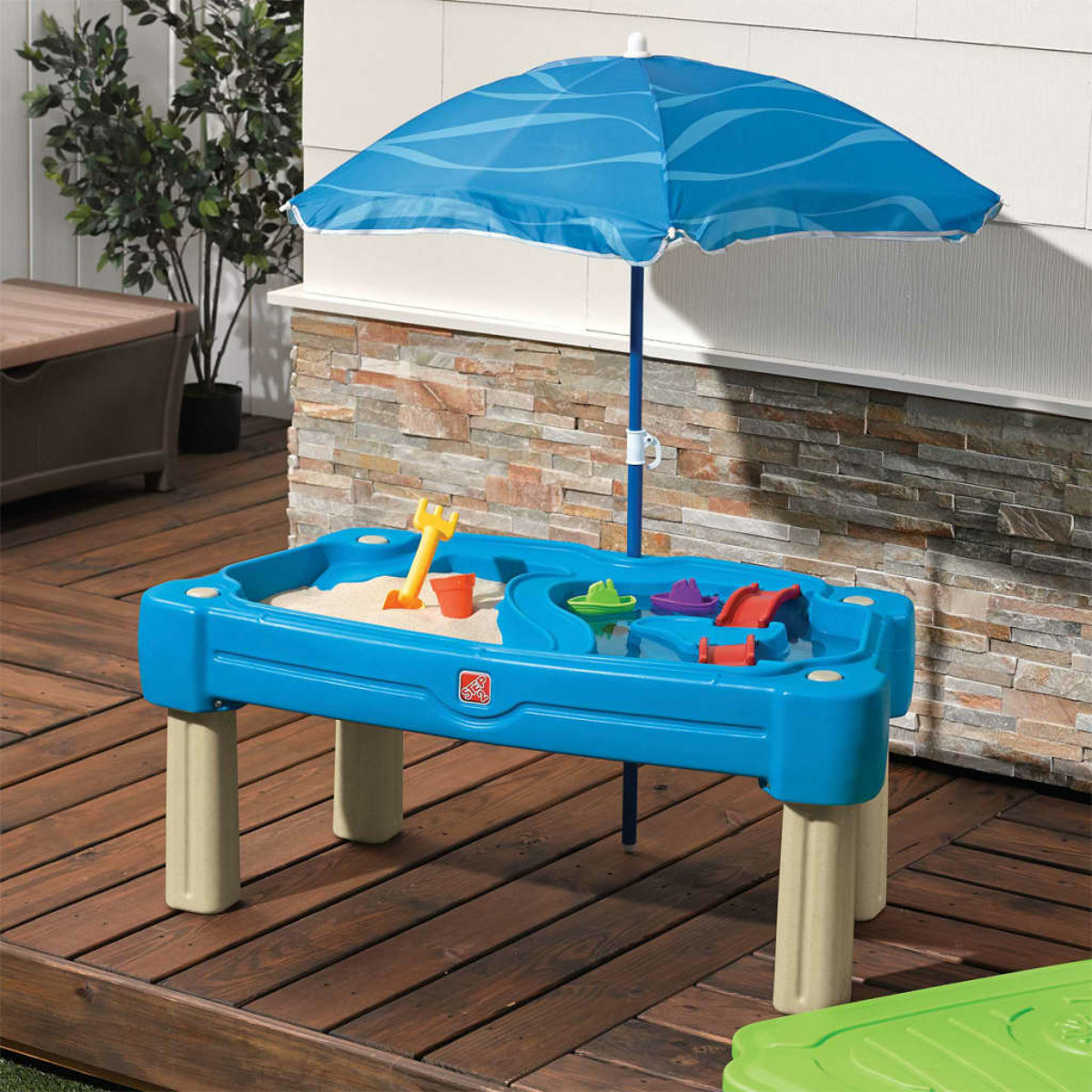 Blau-weißer Sandtisch mit Sonnenschirm, Spielzeug und Sand auf Holzdeck.