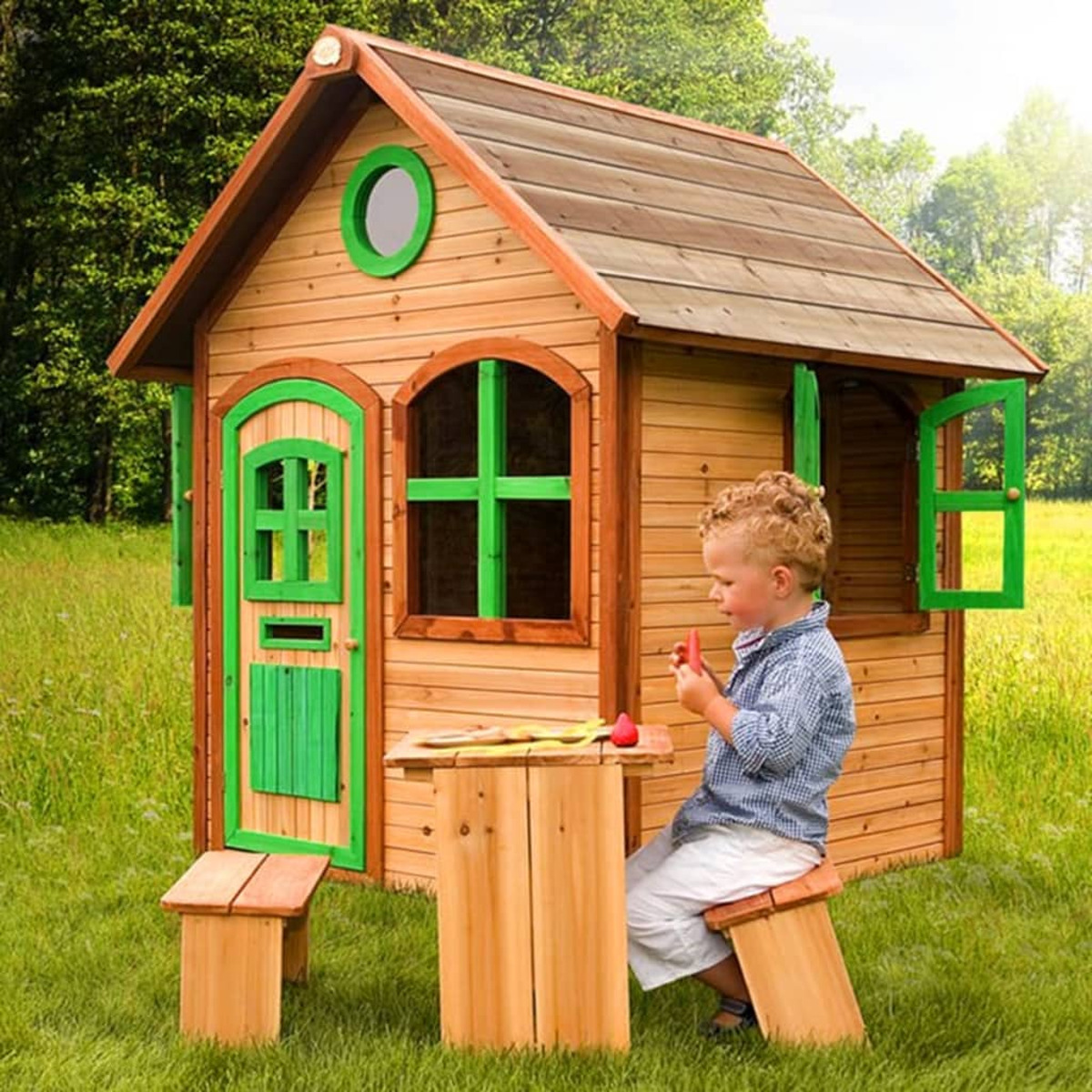 Ein kleiner Junge sitzt an einem Tisch in einem Holzspielhaus und isst. Das Haus ist aus Holz mit grünen Akzenten.