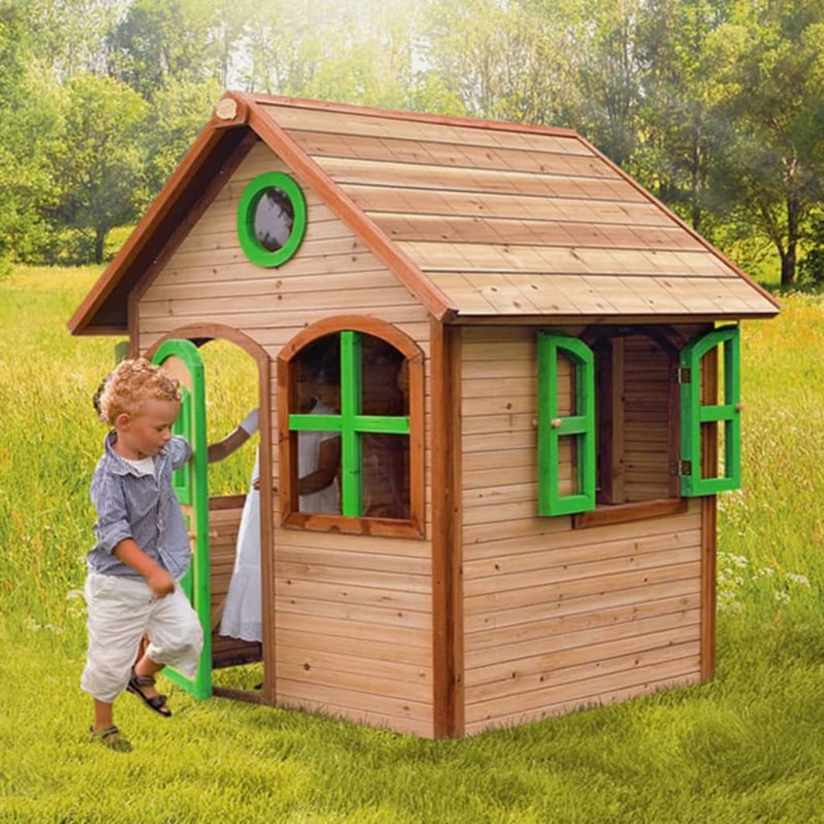 Holzspielhaus mit offener Tür, zwei Kindern, grünen Fensterrahmen und Rasen.