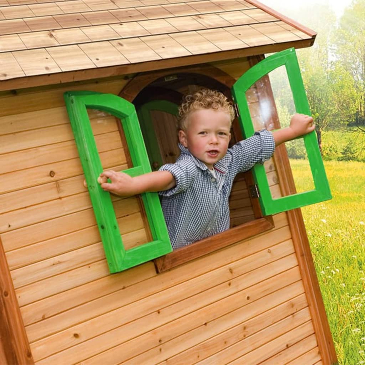 Ein kleiner Junge schaut aus einem Holzspielhausfenster mit grüner Einfassung, Hände am Fensterrahmen.