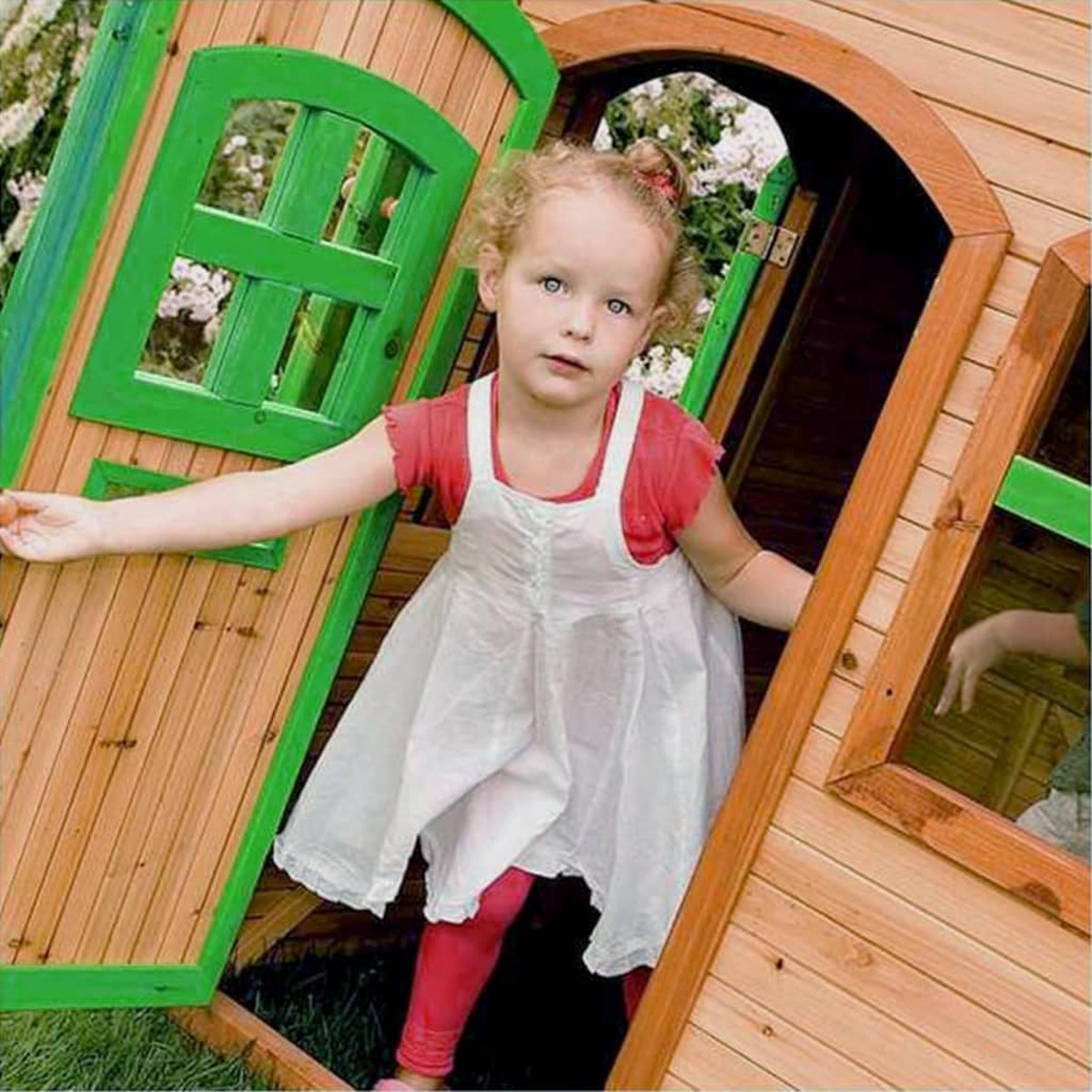 Ein junges Mädchen steht in einer Holzhauseingangstür. Sie trägt ein weißes Kleid und rote Leggings.