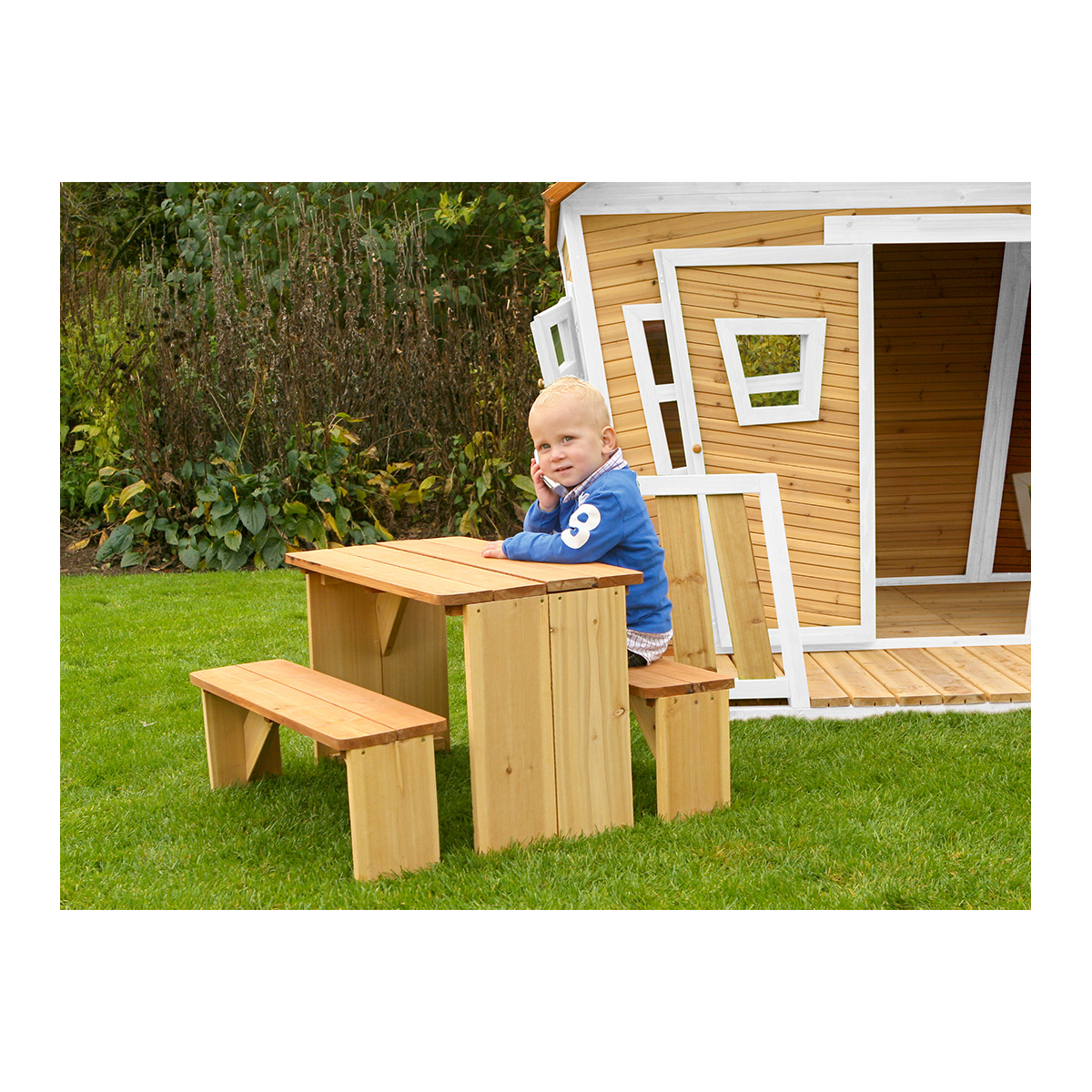 Ein kleiner Junge an einem kleinen Holztisch und einer Bank auf einer Wiese mit Spielhaus.