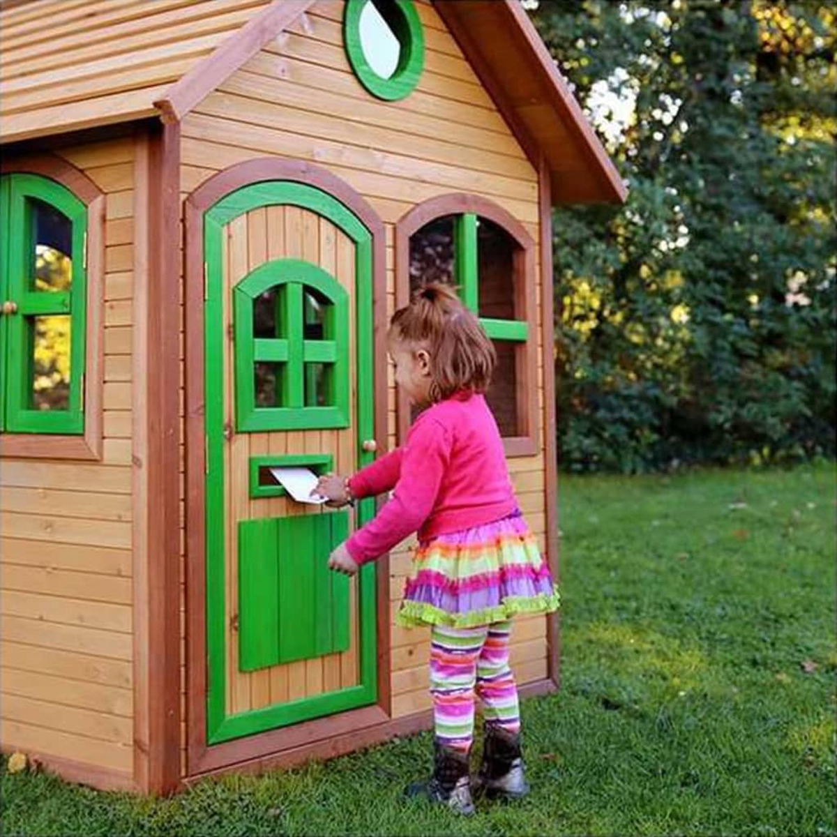 Mädchen wirft Brief in ein Spielhaus mit grünen Akzenten. Draußen, grüne Bäume.