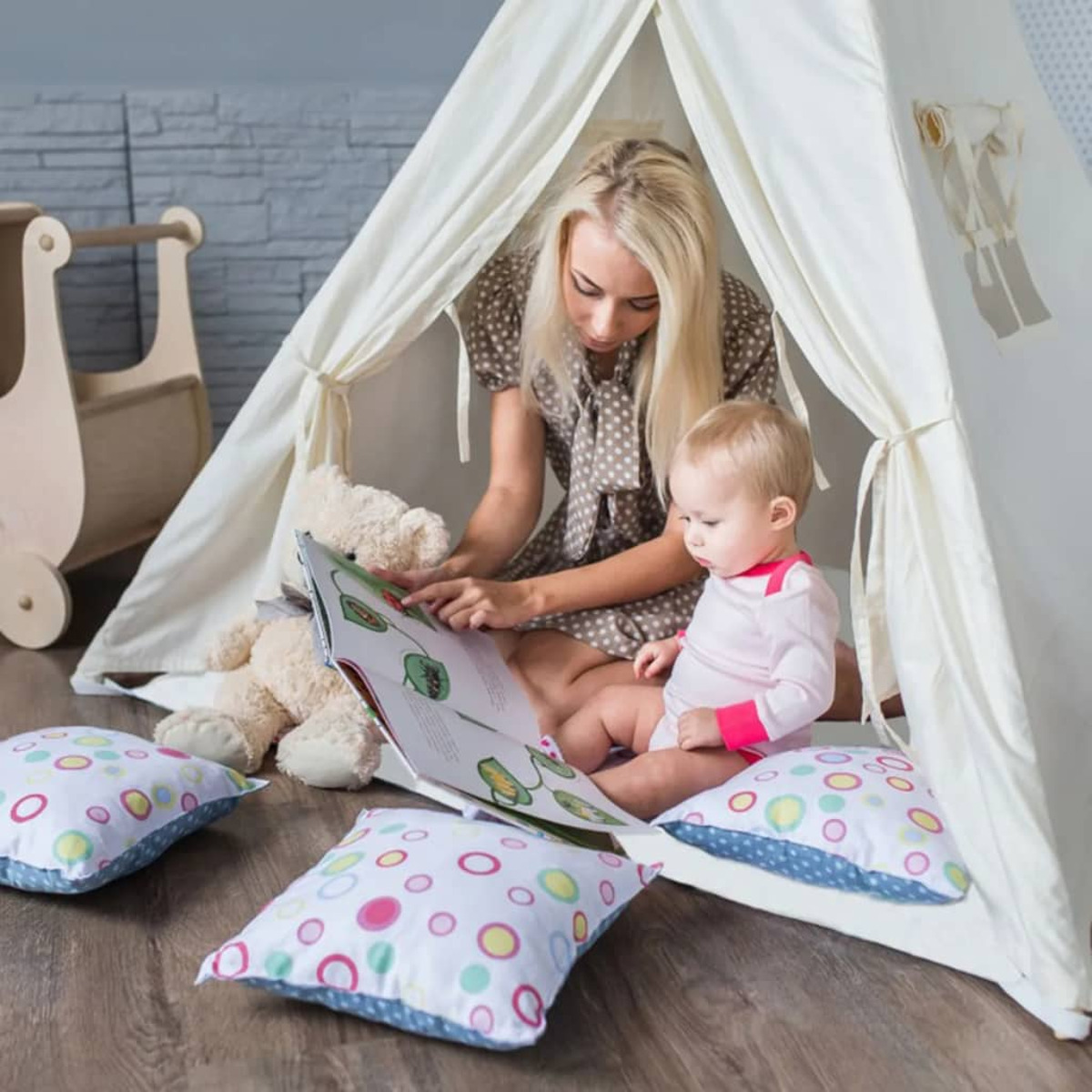 Eine Frau und ein Baby lesen ein Buch in einem weißen Tipi-Zelt mit Kissen und Spielzeug.
