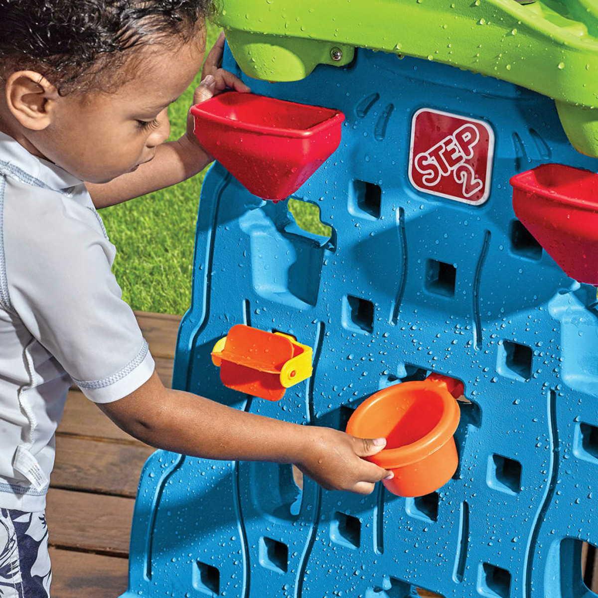 Ein kleiner Junge spielt mit einer Wasserspielwand. Er benutzt einen Becher, um Wasser in die Wand zu gießen.