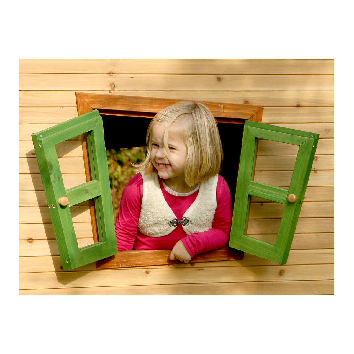 Ein junges Mädchen lächelt aus einem Holzfenster heraus. Das Fenster ist grün und das Holz hellbraun.