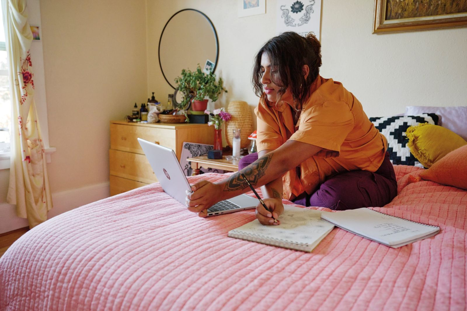 Donna a letto con laptop e quaderno, scrive con la penna.