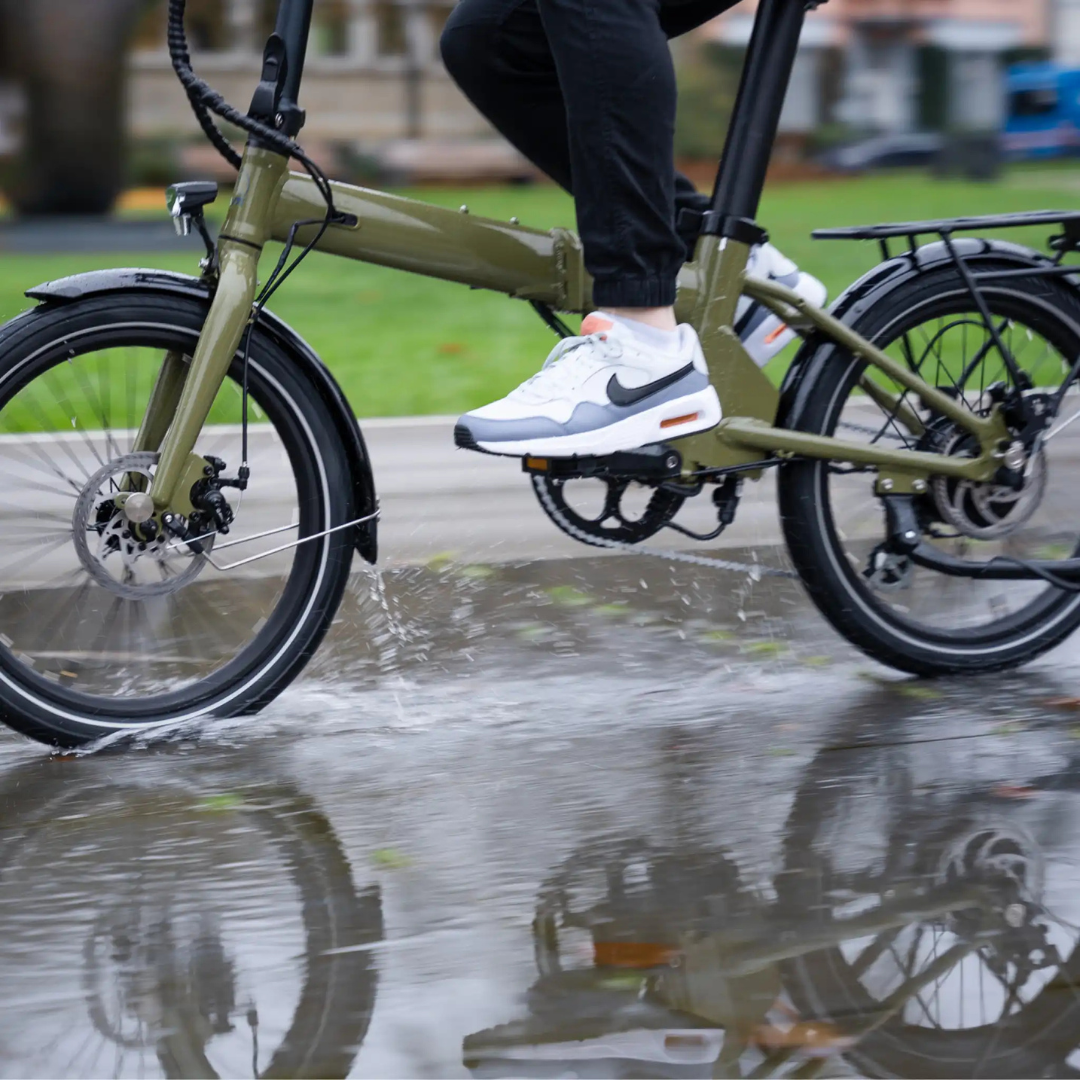 Eine Person fährt mit einem grünen Klapprad durch eine Pfütze und trägt Turnschuhe.