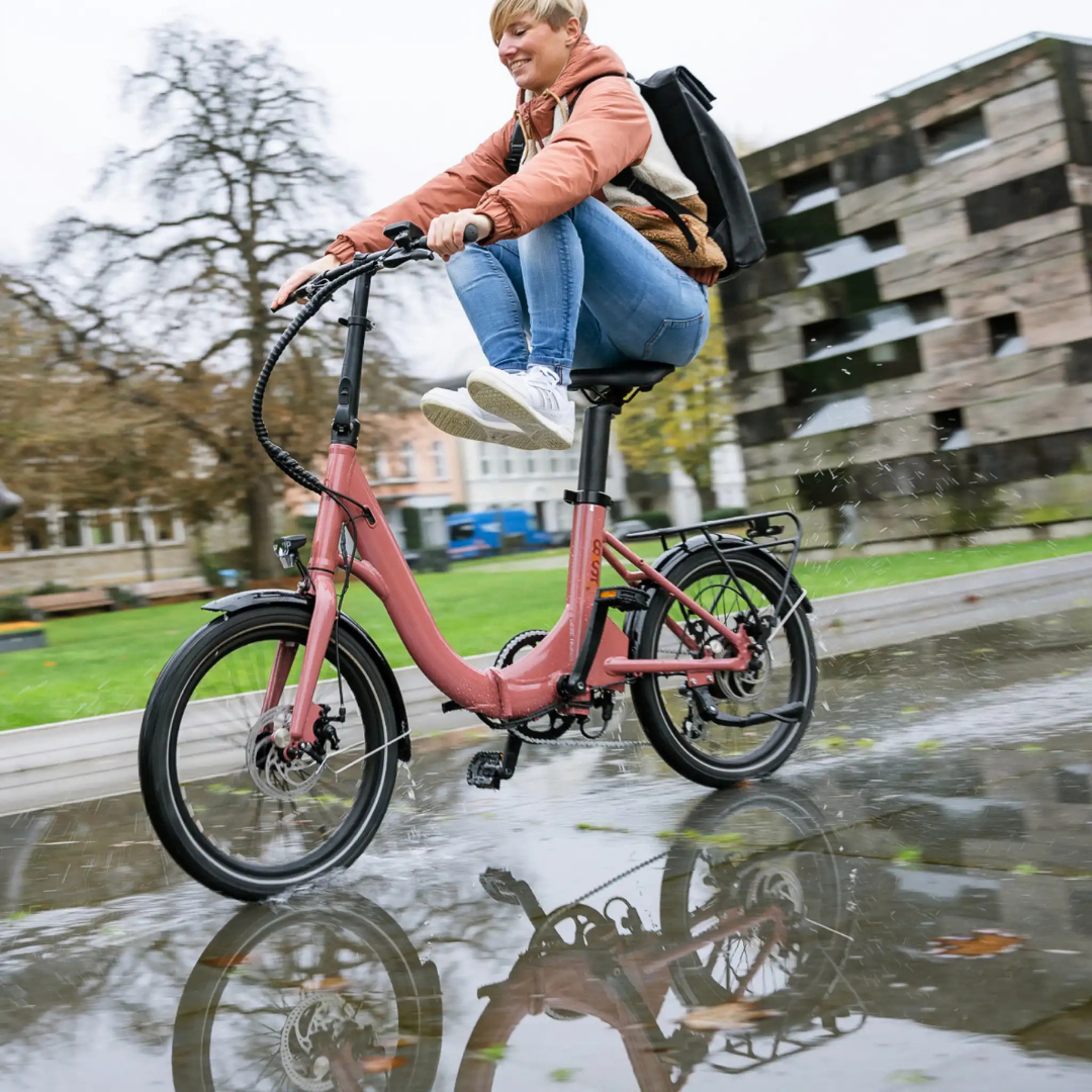 Frau fährt mit einem rosa Faltrad auf einer nassen Straße. Wasserspritzer, Gebäude im Hintergrund.