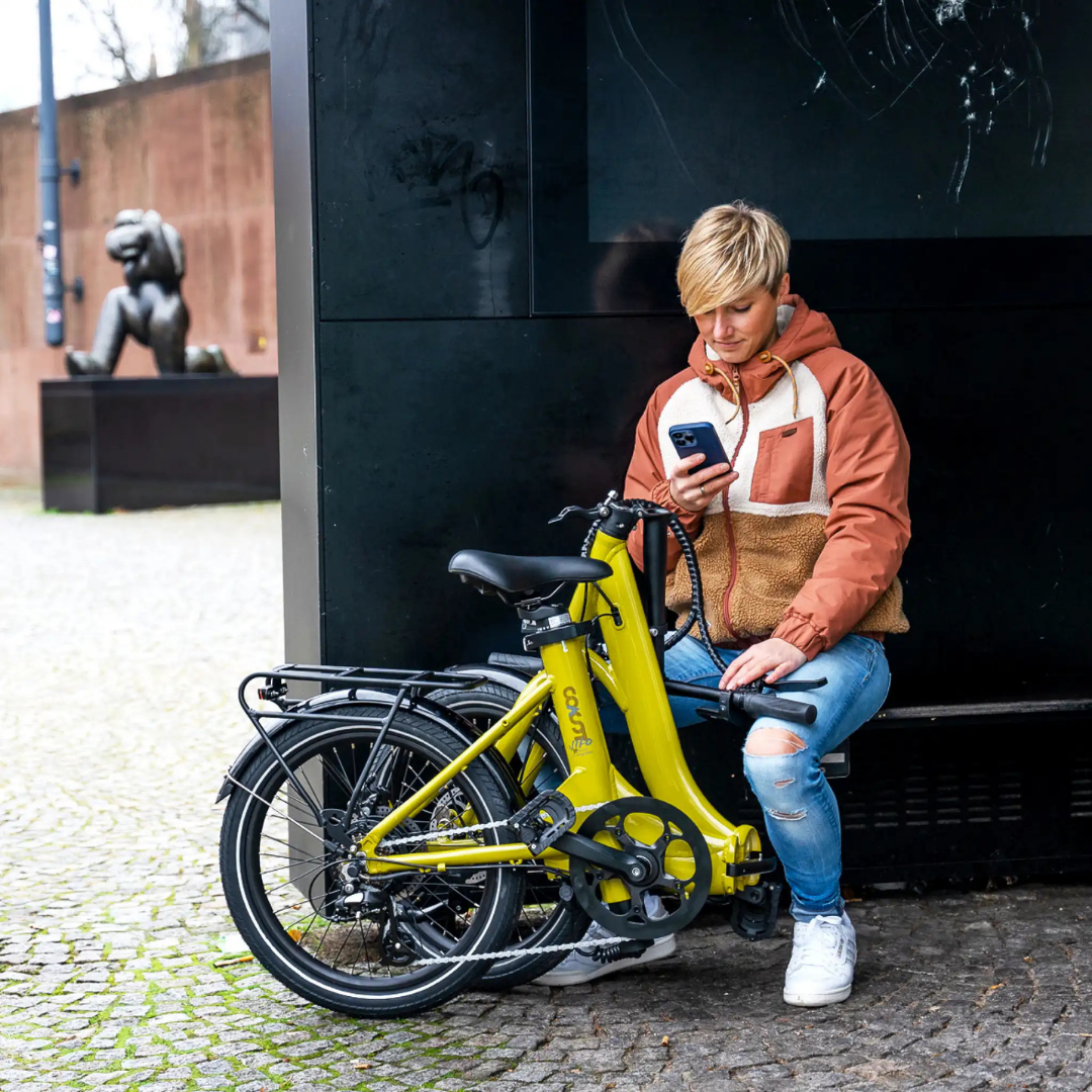 Frau benutzt Telefon, sitzt an gelbem Fahrrad. Hintergrund: Skulptur, Wand, Bank.