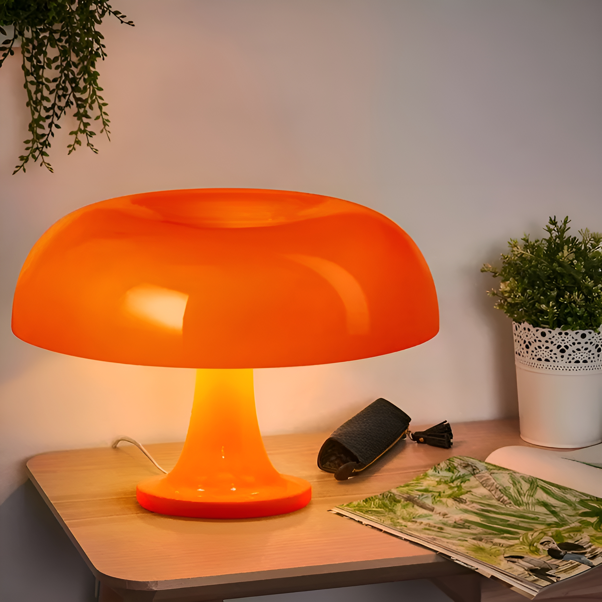 Eine orangefarbene Pilzlampe auf einem Holztisch mit einer Zeitschrift und einer Pflanze. Die Wand ist weiß.
