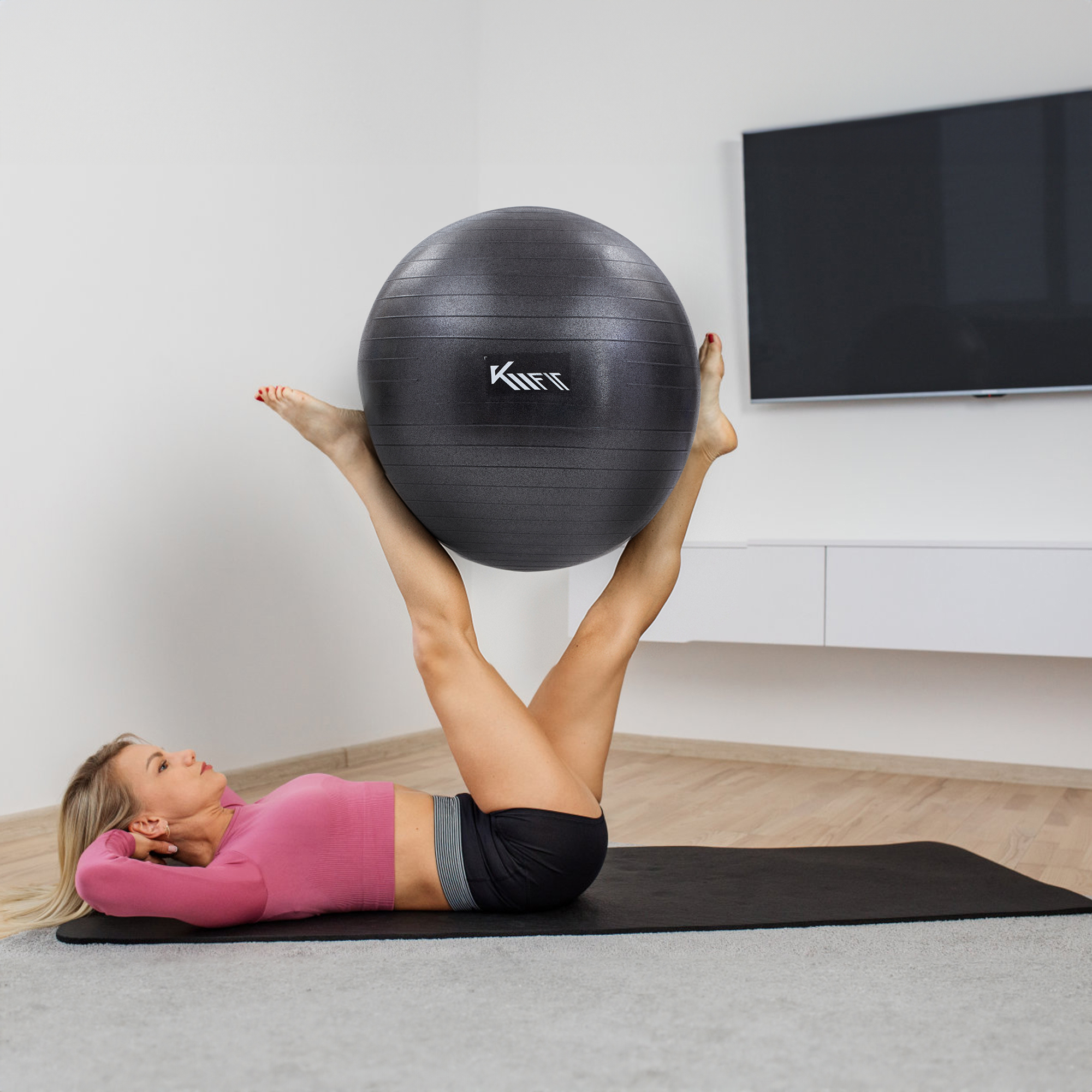 Eine Frau liegt auf einer Matte und hält einen schwarzen Gymnastikball zwischen ihren Beinen. TV im Hintergrund.