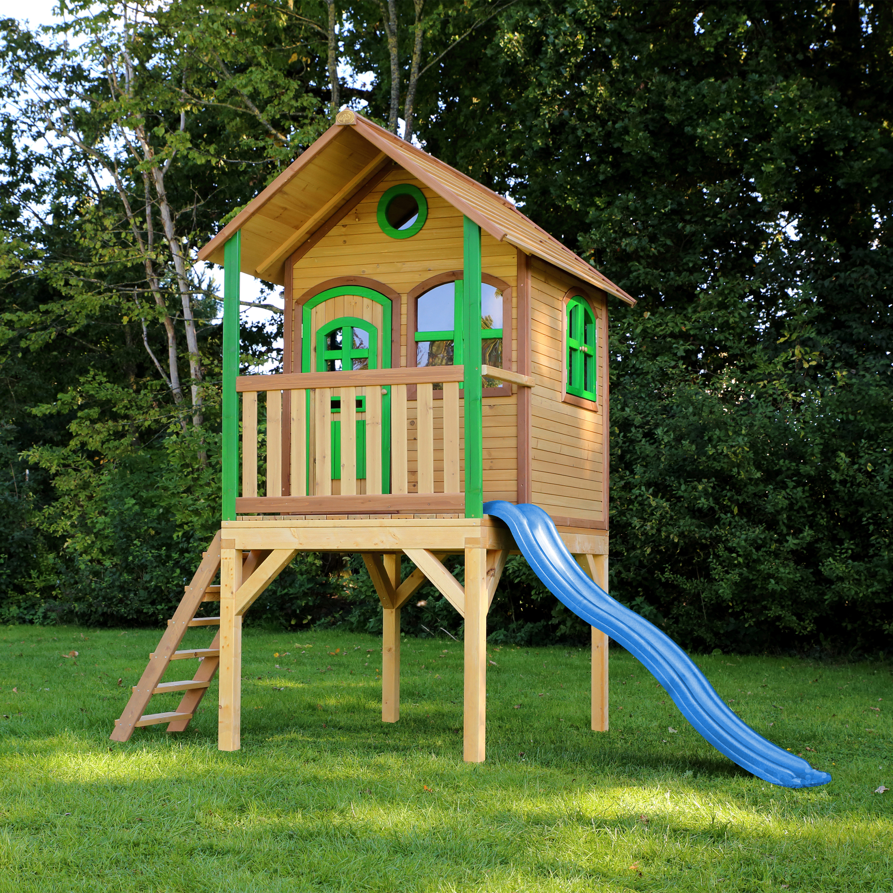 Baumhaus aus Holz mit grünen Akzenten, einer Rutsche und einer Leiter. In einem grasbewachsenen Bereich.