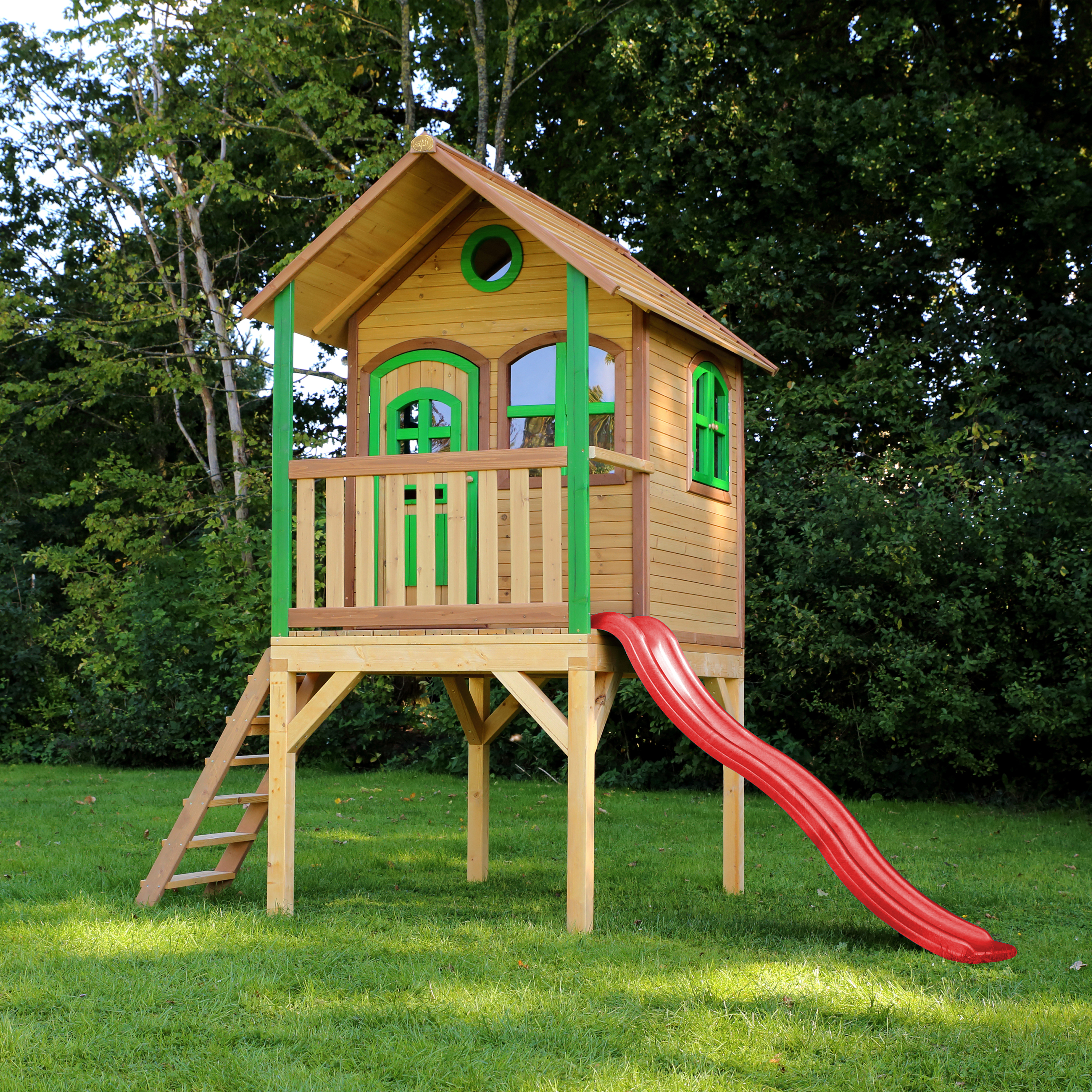 Holzspielhaus auf einer Wiese. Es hat eine Leiter, Rutsche und grüne Akzente an Fenstern und Balkon.