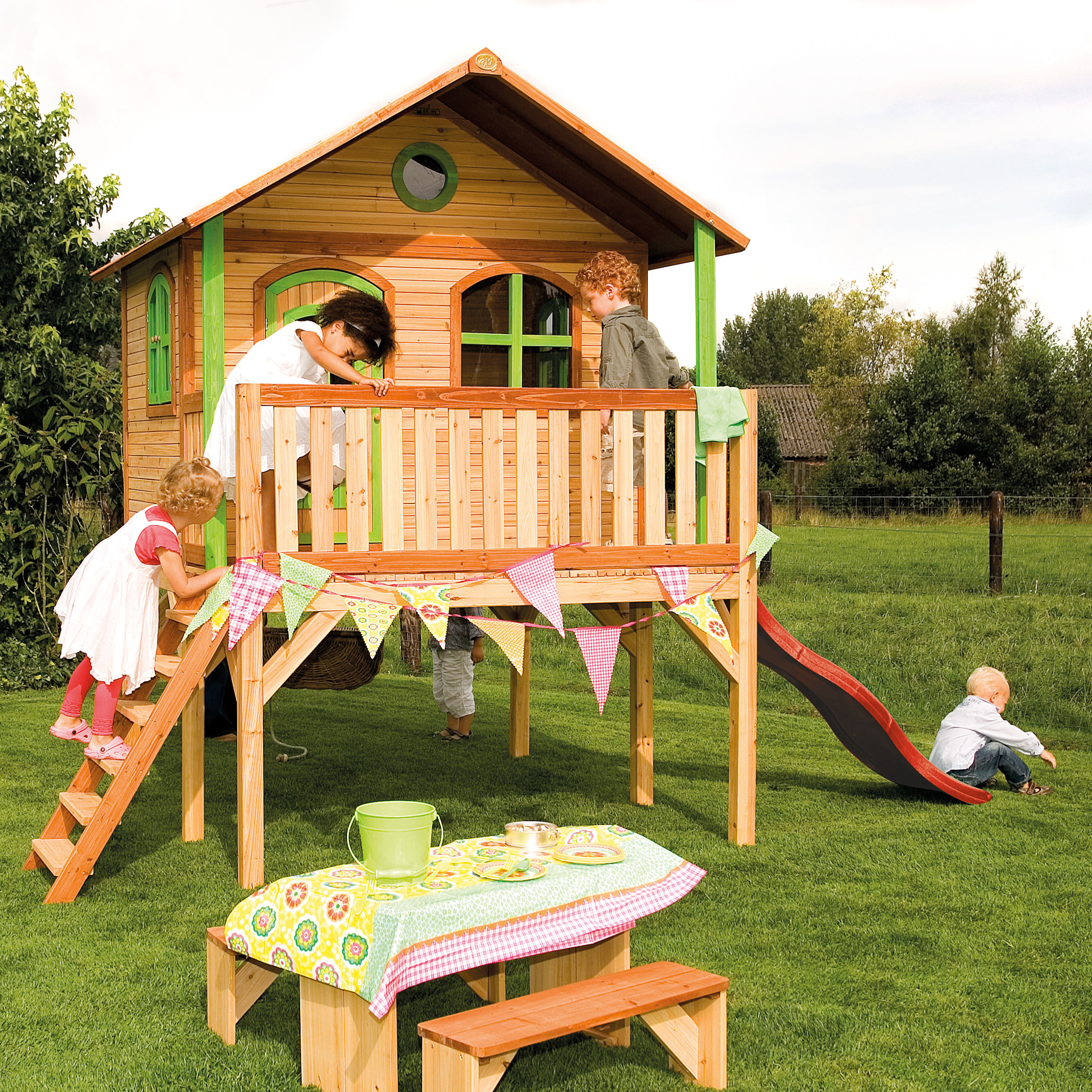Kinder spielen auf einem Holzspielhaus mit Rutsche. Ein Picknicktisch steht im Gras.