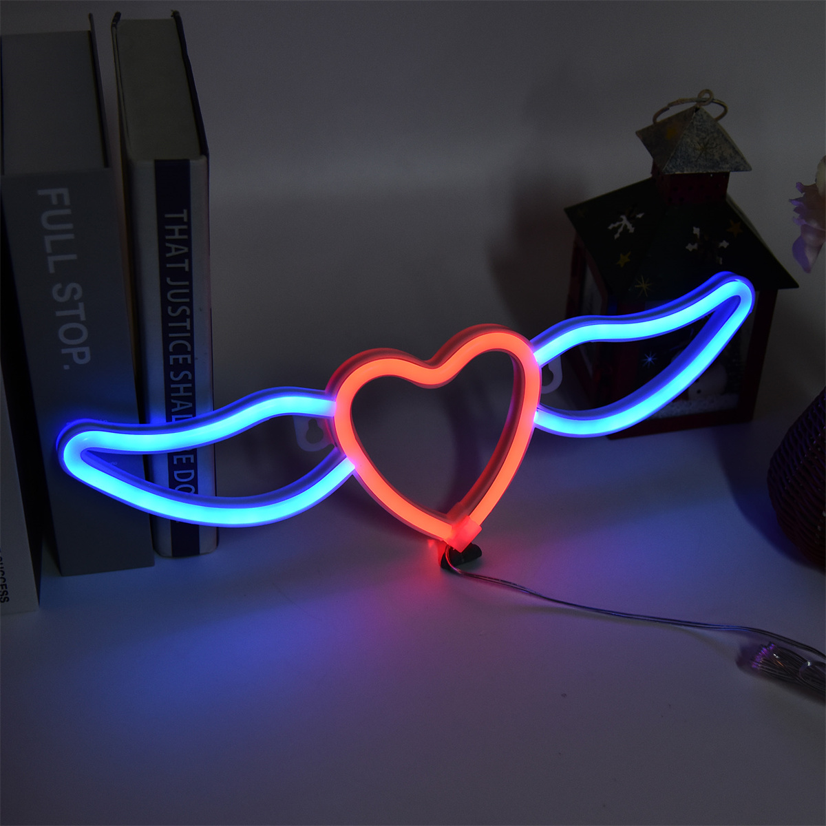 Neon-Herzförmiges Licht mit Flügeln. Das Herz ist rot, die Flügel blau. Auf einem Regal mit Büchern platziert.